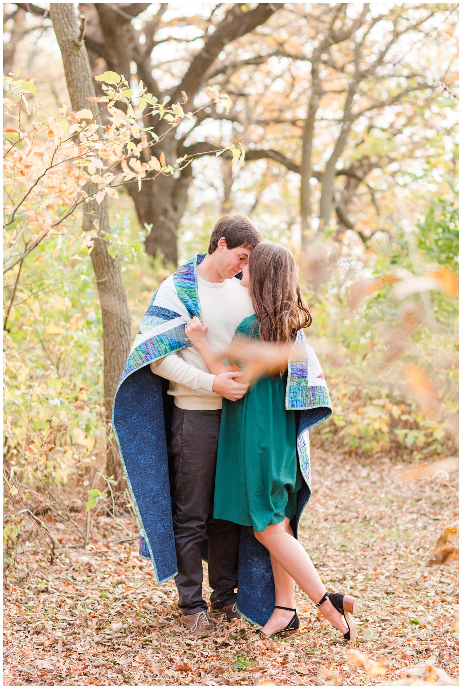 Fall in Iowa, Jenna wearing an emerald green dress embraces Brady while both wrapped in a quilt in the middle of an autumn path at Lost Island Nature Center | CB Studio