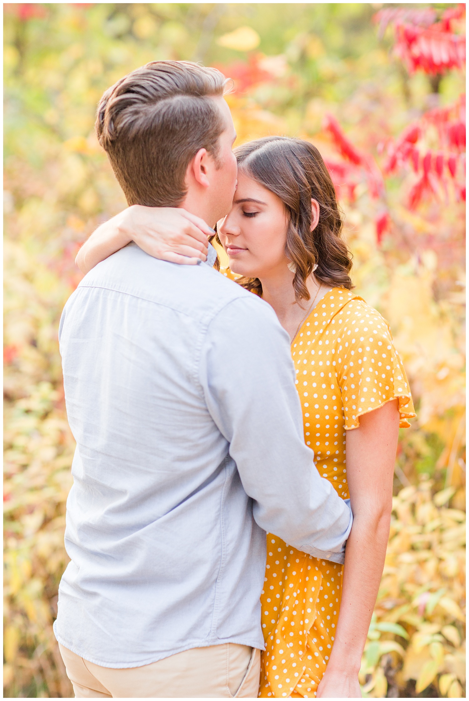 Jadi wearing a yellow flowing dress looks down as Luke kisses her forehead while embracing together in a grassy field in Iowa during the fall for engagement photos | CB Studio