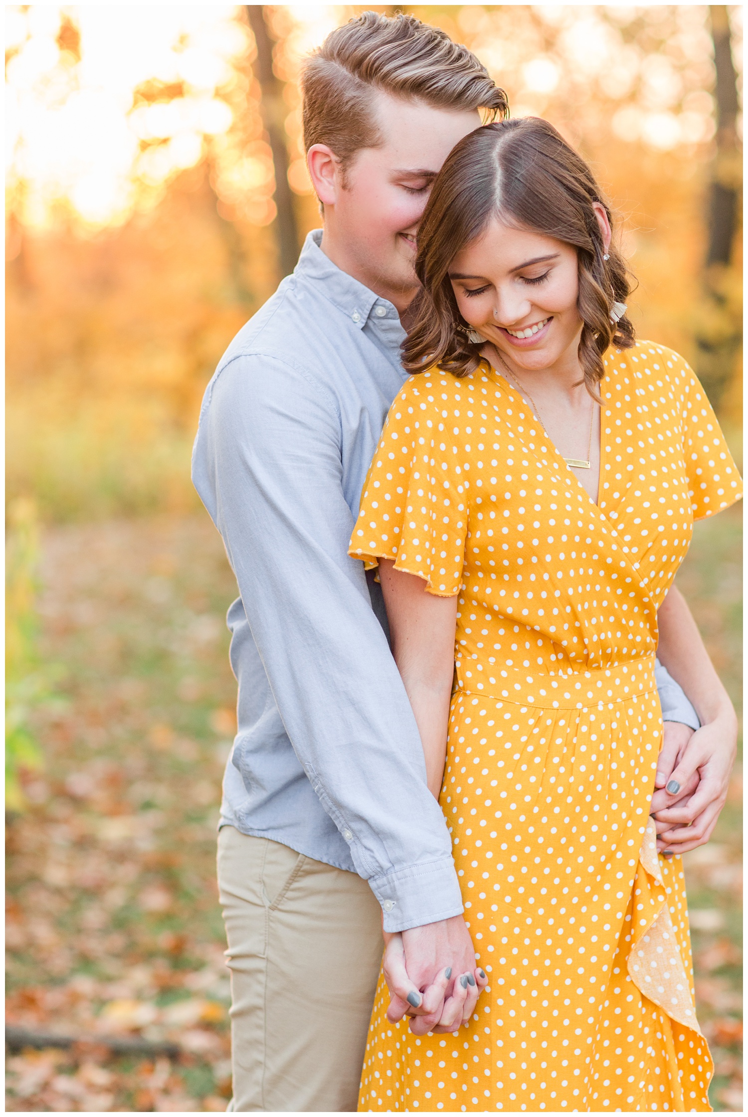 Jadi wearing a yellow flowing dress smiles as Luke whispers into her ear while embracing together in a grassy field in Iowa during the fall for engagement photos | CB Studio