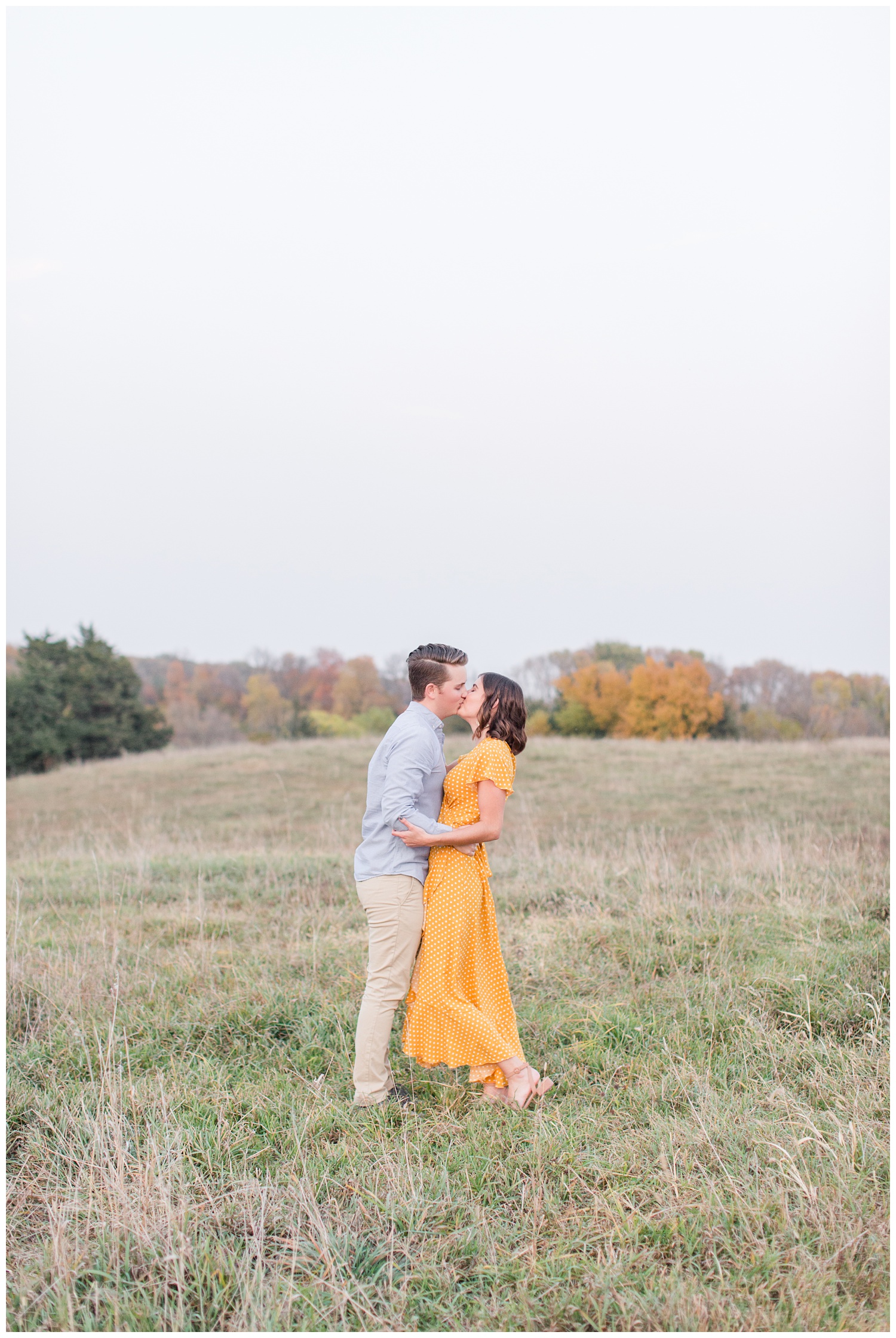 Jadi wearing a yellow flowing dress kisses Luke while embracing together in a grassy field in Iowa during the fall for engagement photos | CB Studio