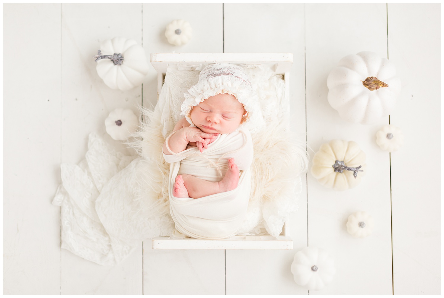 Newborn baby girl wrapped in white snuggled on a white baby bed surrounded by white pumpkins in a light and bright fall inspired newborn pose setup | CB Studio