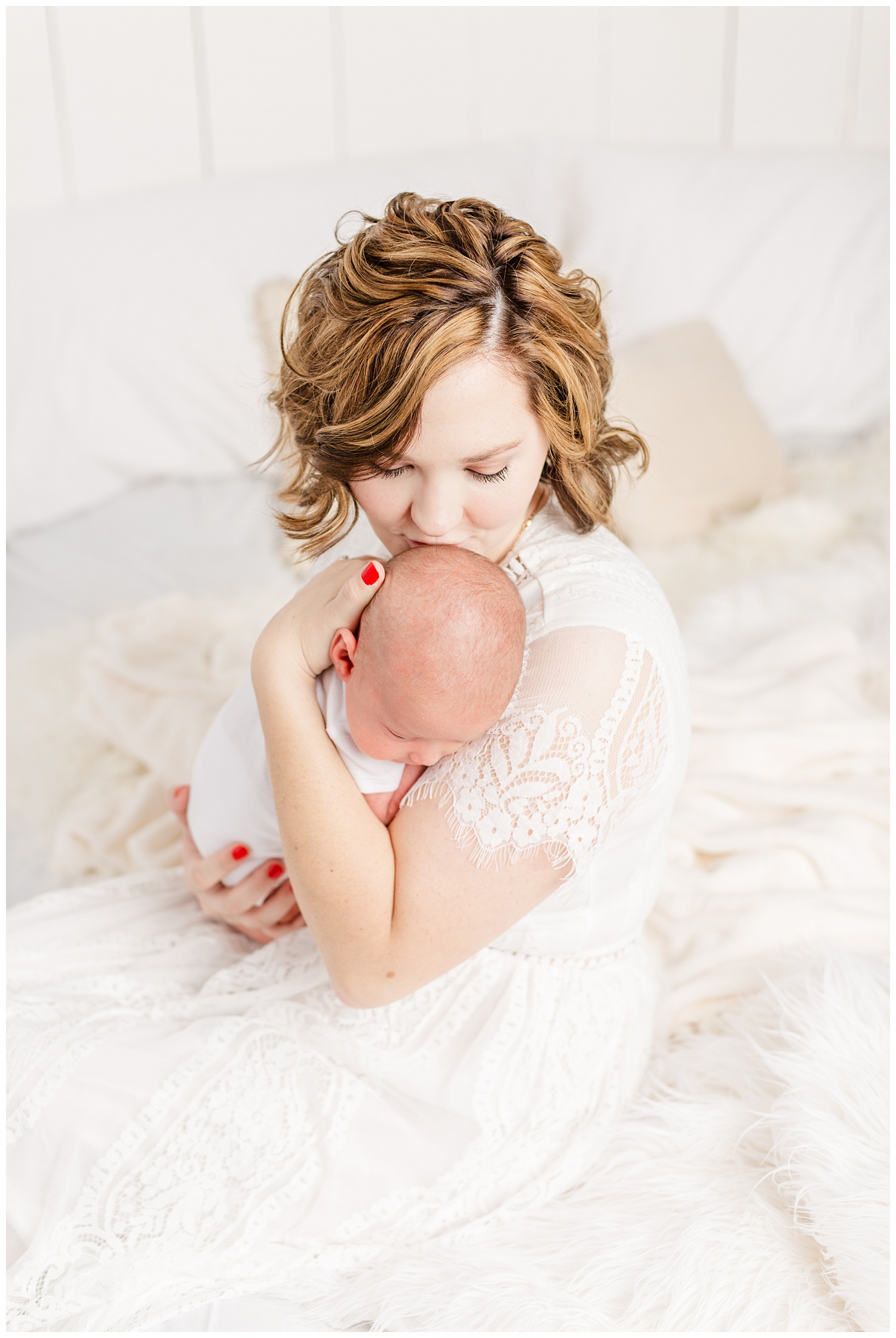 Bree dressed in white lace sits on her bed and gently kisses her new baby boy | CB Studio