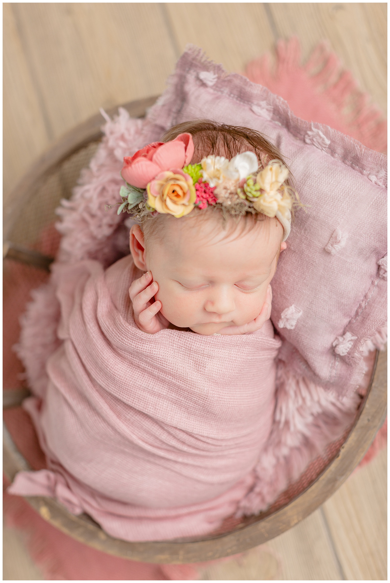 Newborn baby girl wrapped in a mauve colored stretch fabric snuggled in a basket bowl wearing a garden floral headband. | CB Studio