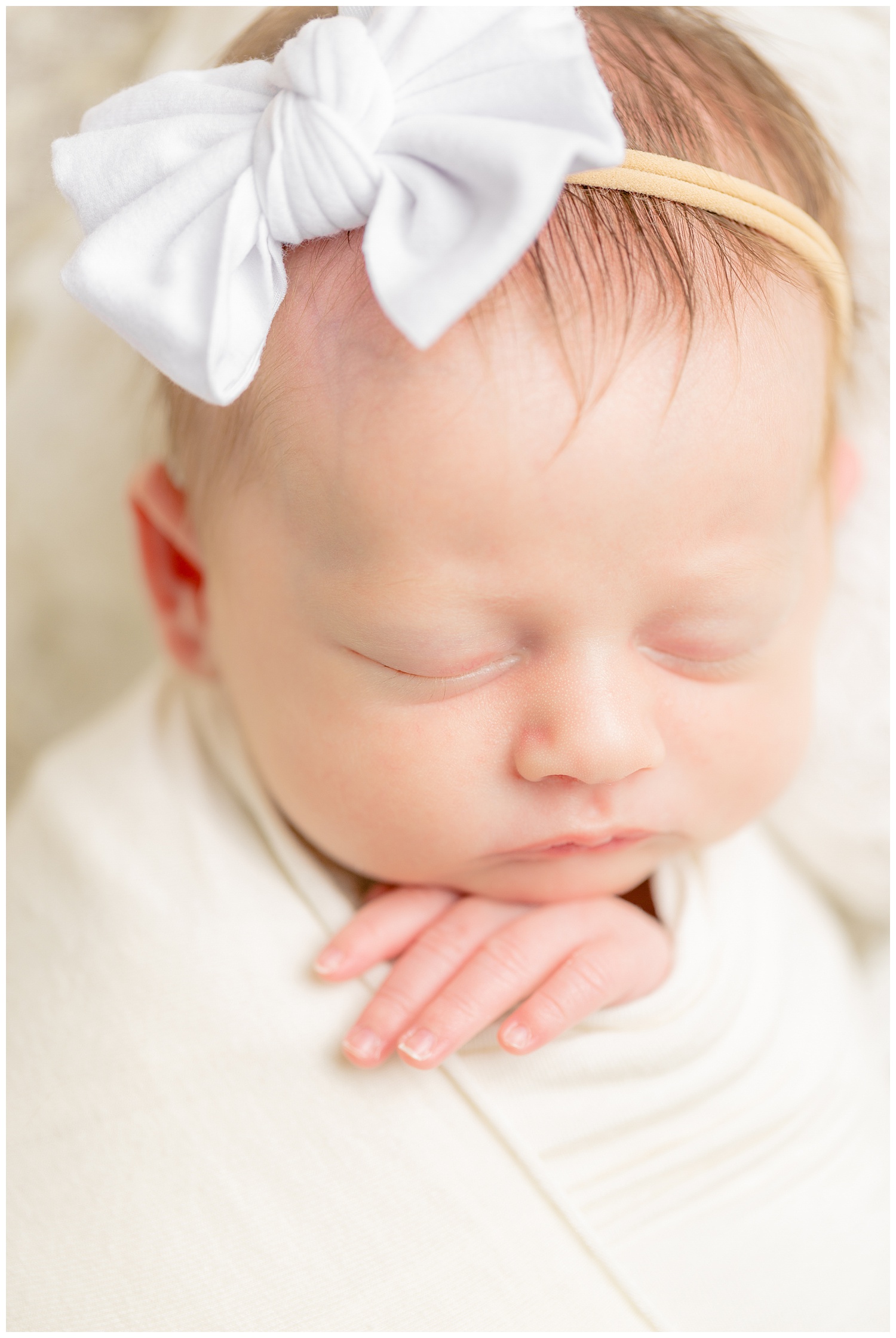 Newborn baby girl wrapped in ivory stretch fabric resting her chin gently on her hand while wearing a white bow headband. | CB Studio