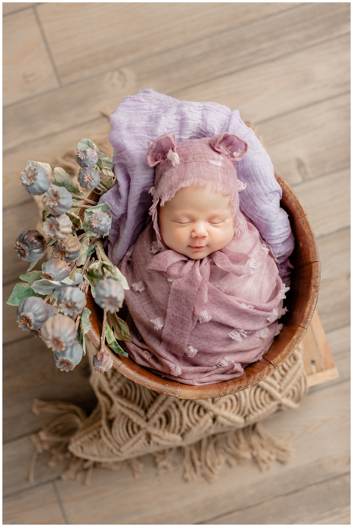 Newborn baby girl Remi swaddled in a mauve textured wrap and bear ear bonnet lying in a basket full of flowers | CB Studio