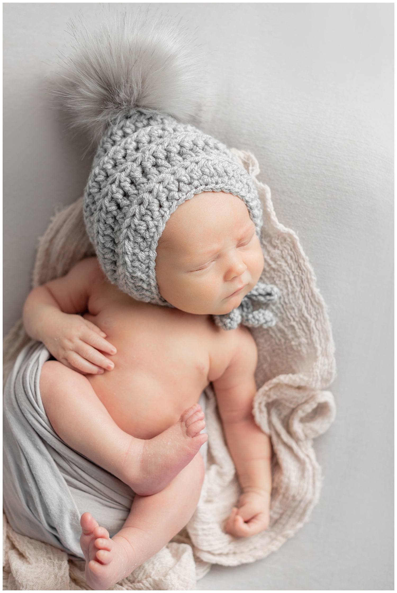 Baby Braxton sleeps soundly on a grey fabric background wearing a grey knit pom bonnet | CB Studio