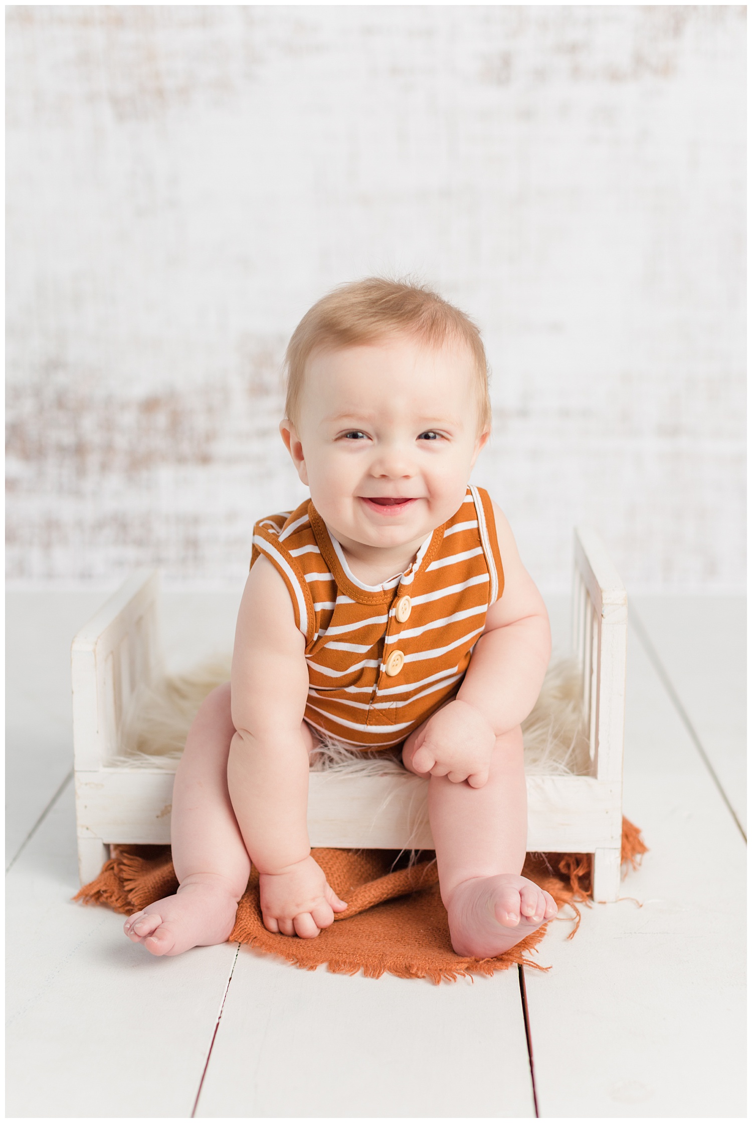 Baby Jackson sitting on a little white bed wearing a camel colored romper | CB Studio
