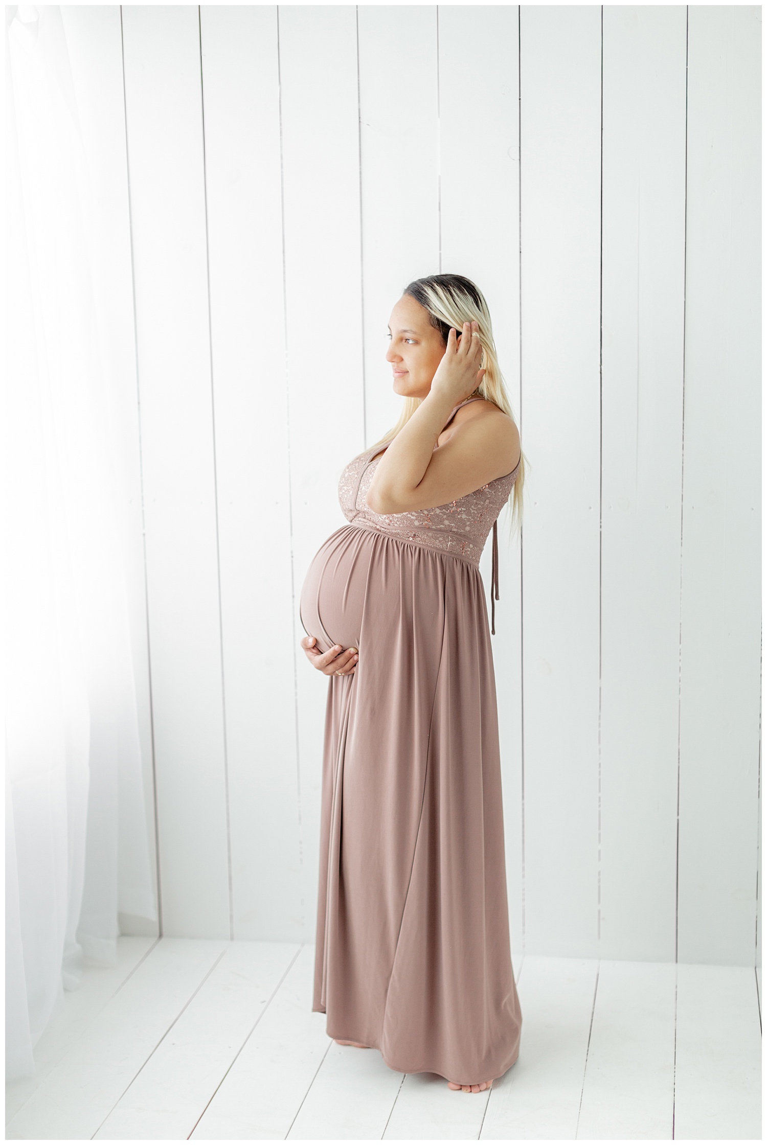 Karen patiently waits for the arrival of her baby girl while standing in a white wood room wearing a blush maxi dress and holding her baby bump | CB Studio
