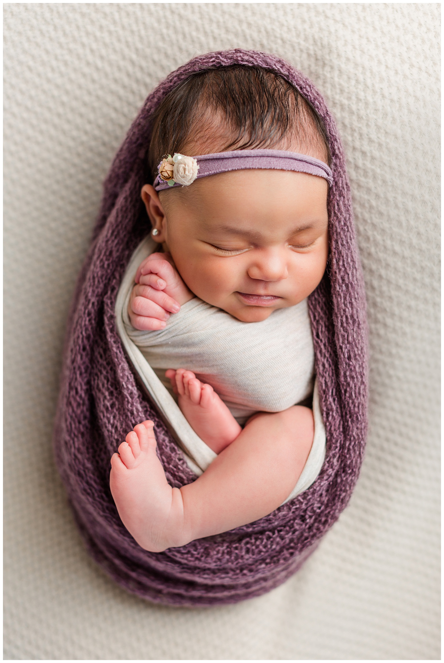 Newborn baby Alyssa wrapped up in a puprle knit wrap with her hands folded in prayer and smiling | CB Studio