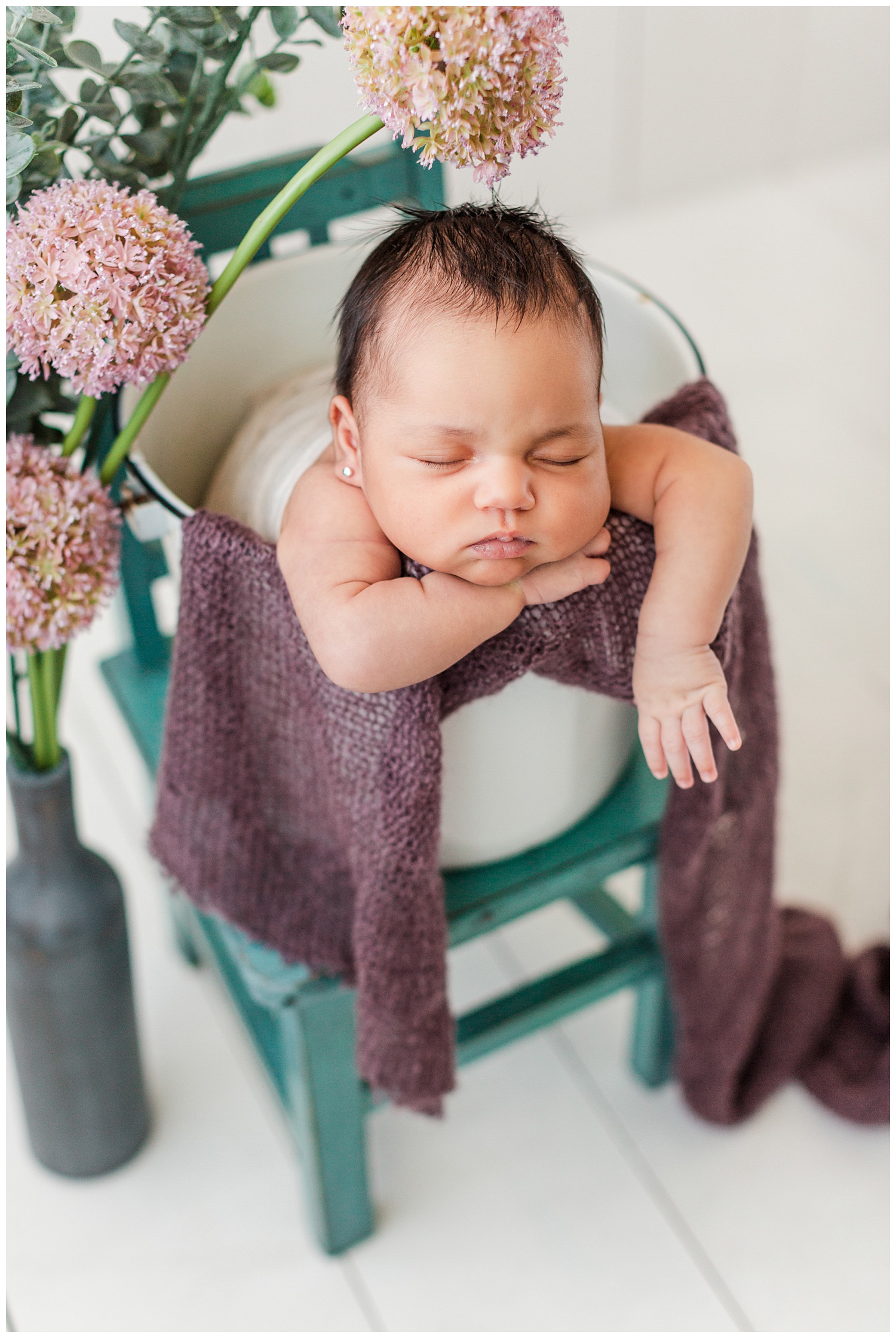 Newborn baby Alyssa snuggled in a white bucket resting on a teal chair with beautiful pink florals surrounding her | CB Studio