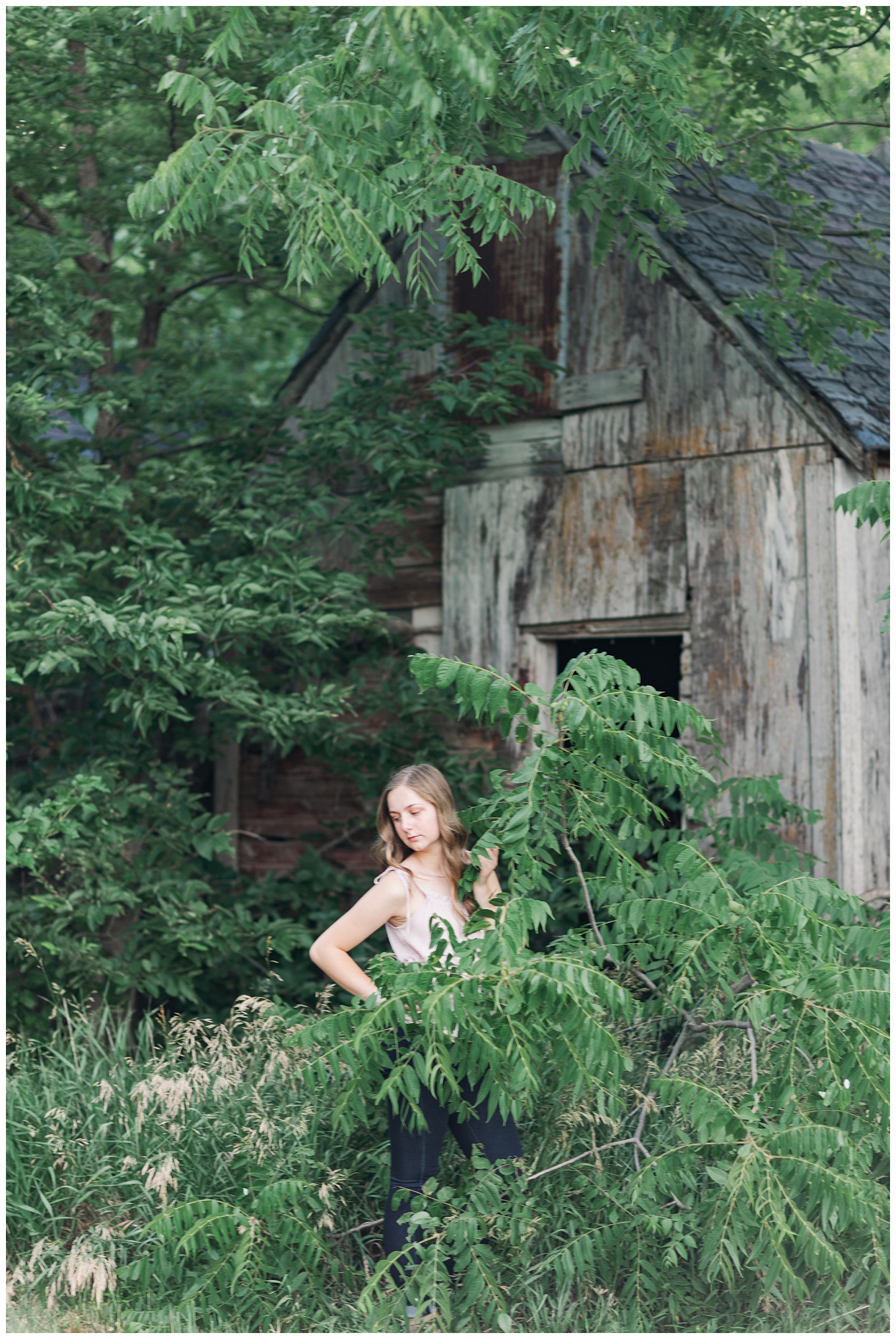 Senior Taylor looks down as she is surrounded by green foliage with a wood barn in the background | CB Studio