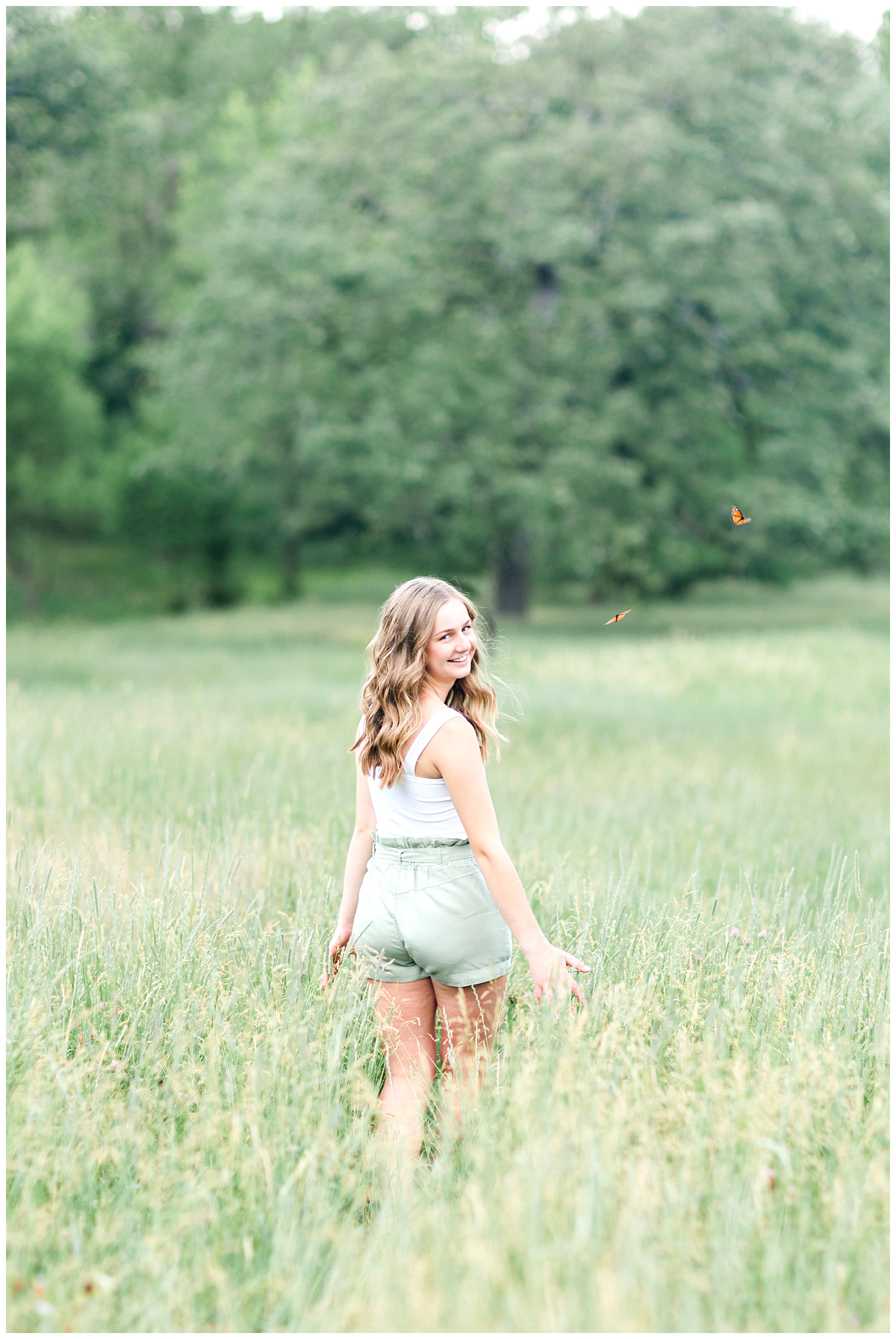 Senior Taylor walking away in a grassy field with butterflies flying around her | CB Studio