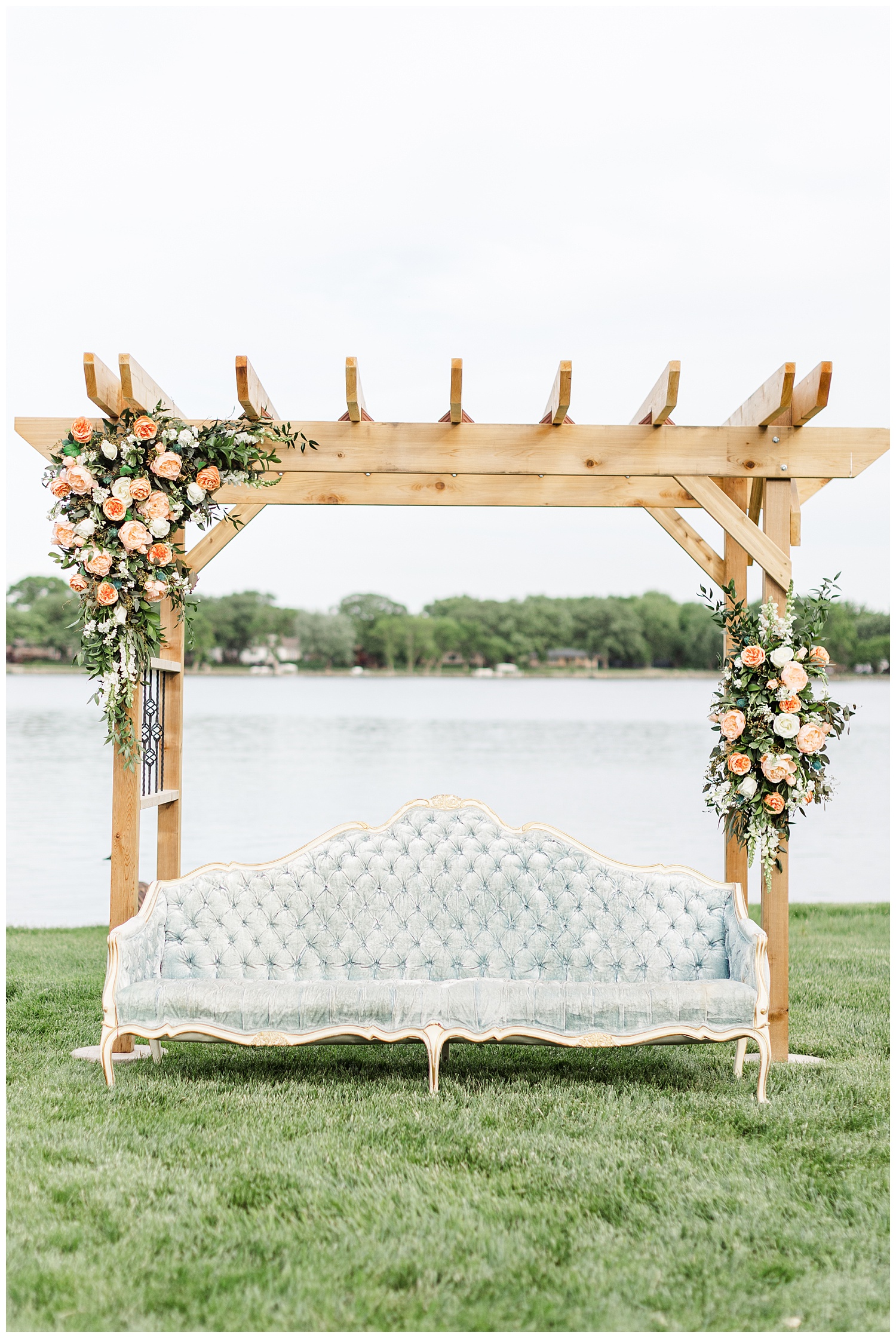 Wedding arch adorned with peach garden roses and accompanied by a dusty blue victorian couch with a beautiful lake background | CB Studio