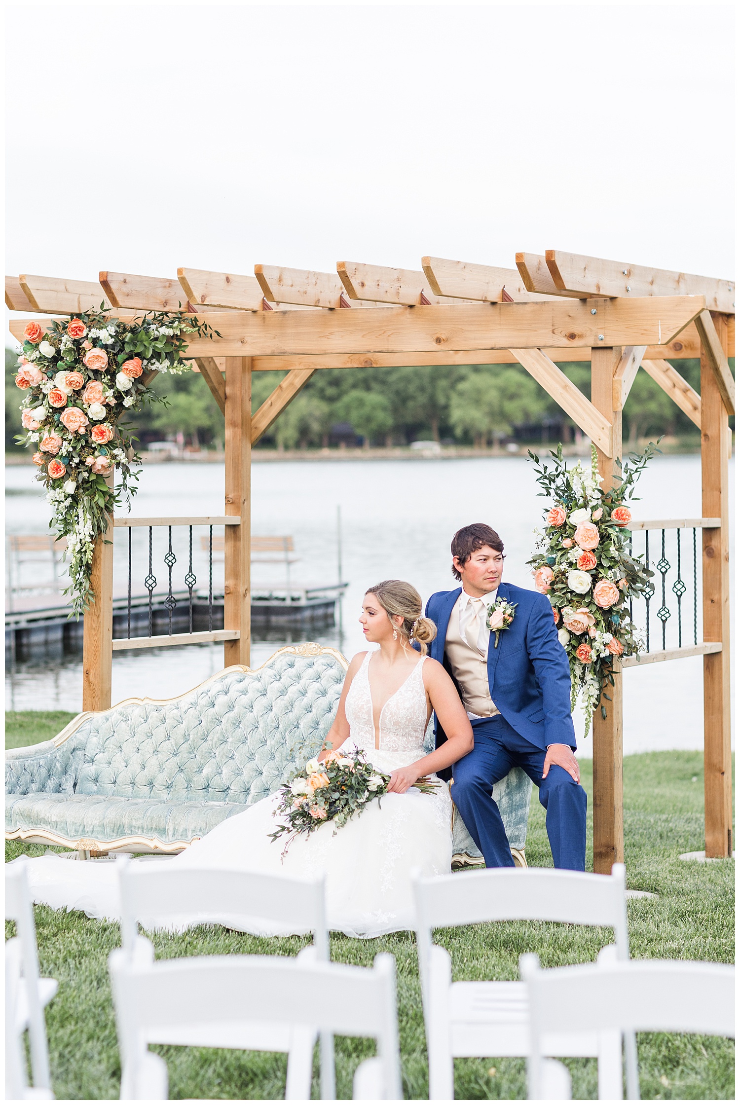 Bride and groom sit on a dusty blue victorian couch under a wooden wedding arch adorned with peach garden roses | CB Studio