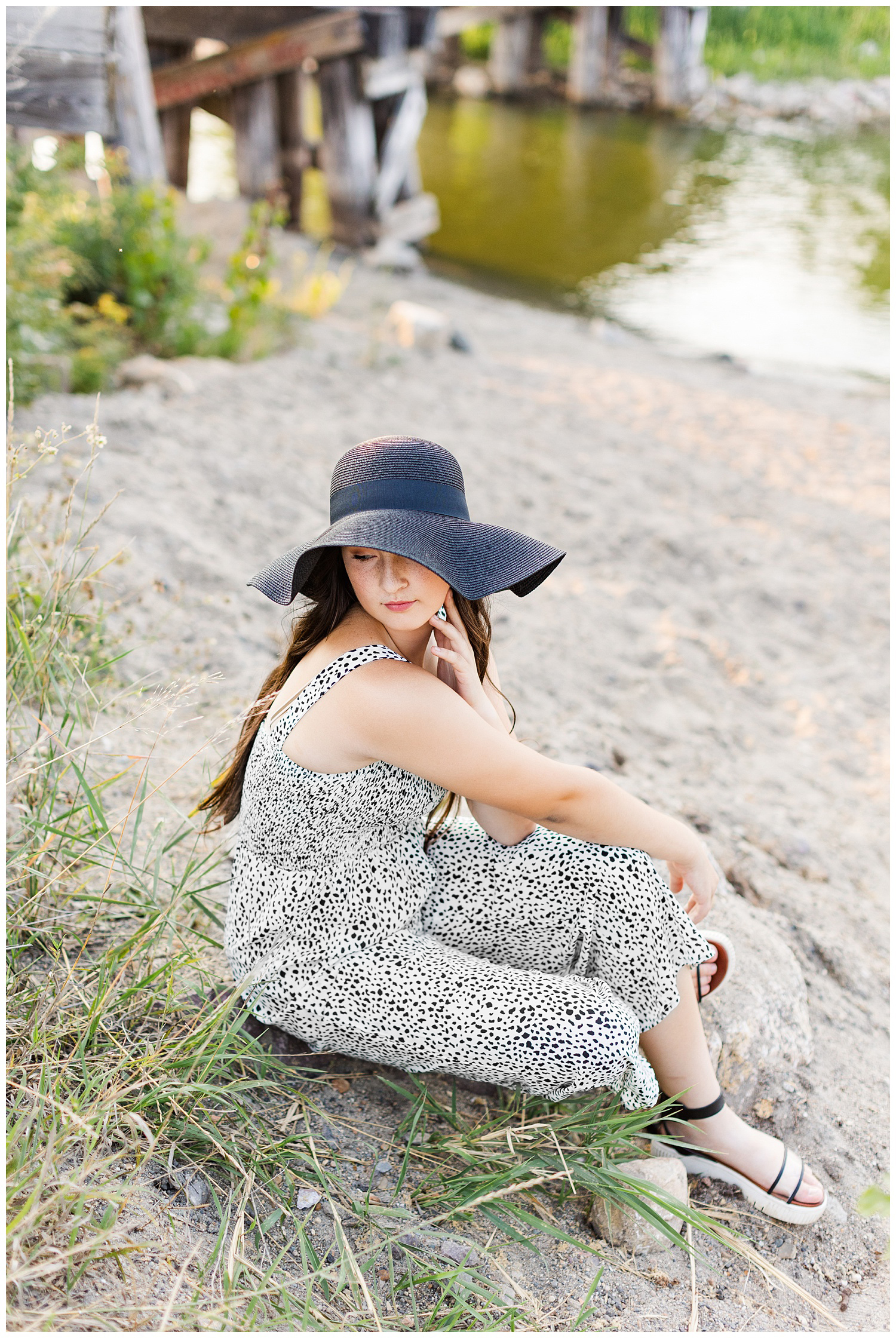Laney wearing a black and white jumpsuit and black sun hat looks over her shoulder while sitting on the shores of Five Island Lake in Emmetsburg | CB Studio