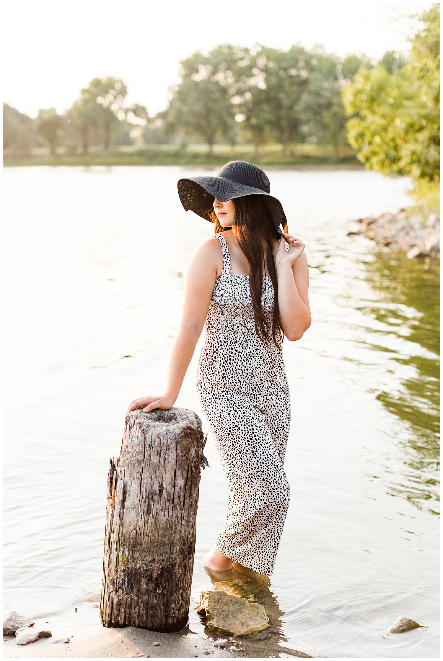 Laney wearing a black and white jumpsuit and black sun hat looks over her shoulder while standing in the water of Five Island Lake in Emmetsburg | CB Studio