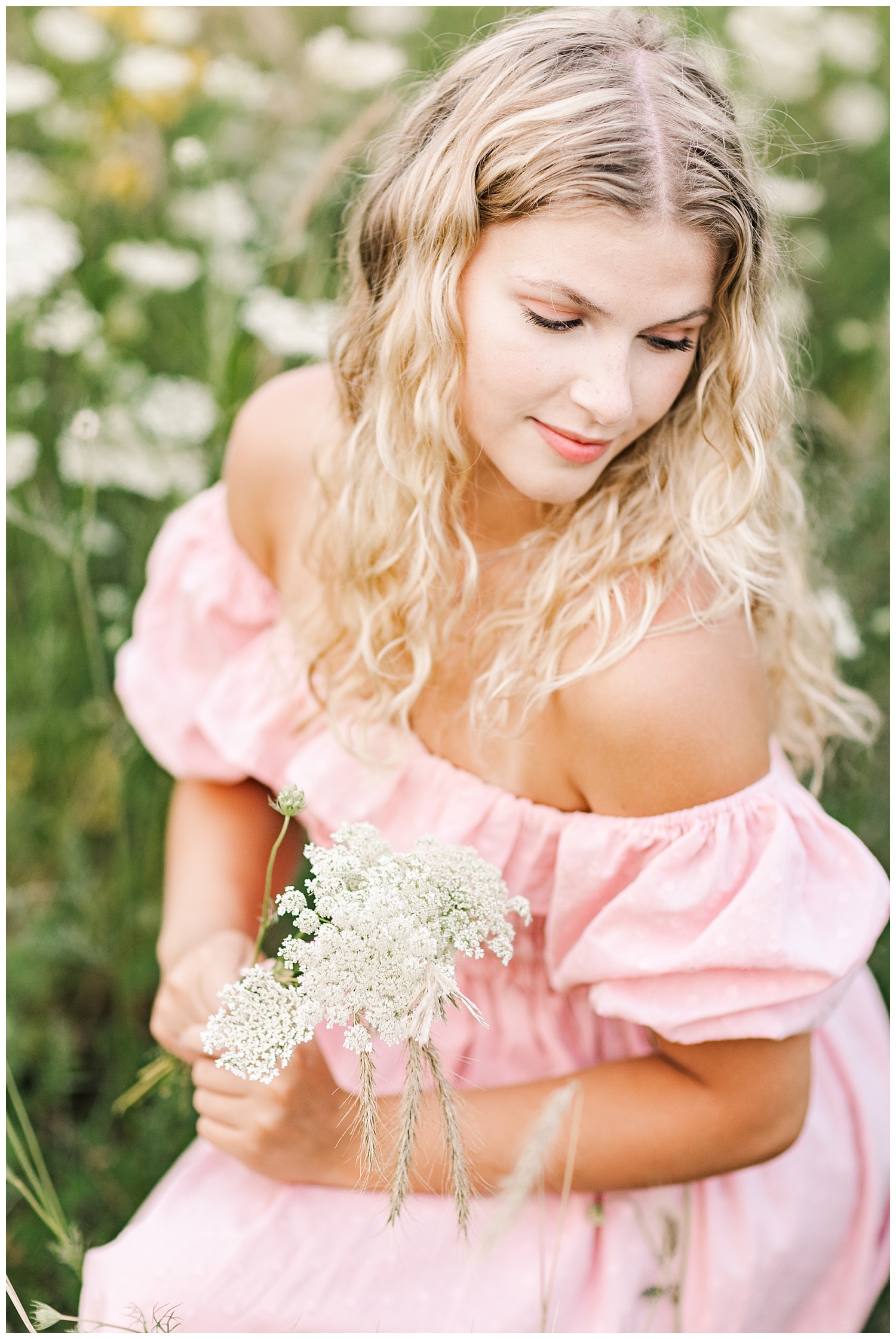 Senior Rachel wearing a light pink off the shoulder dress crouches down in a wildflower field | CB Studio