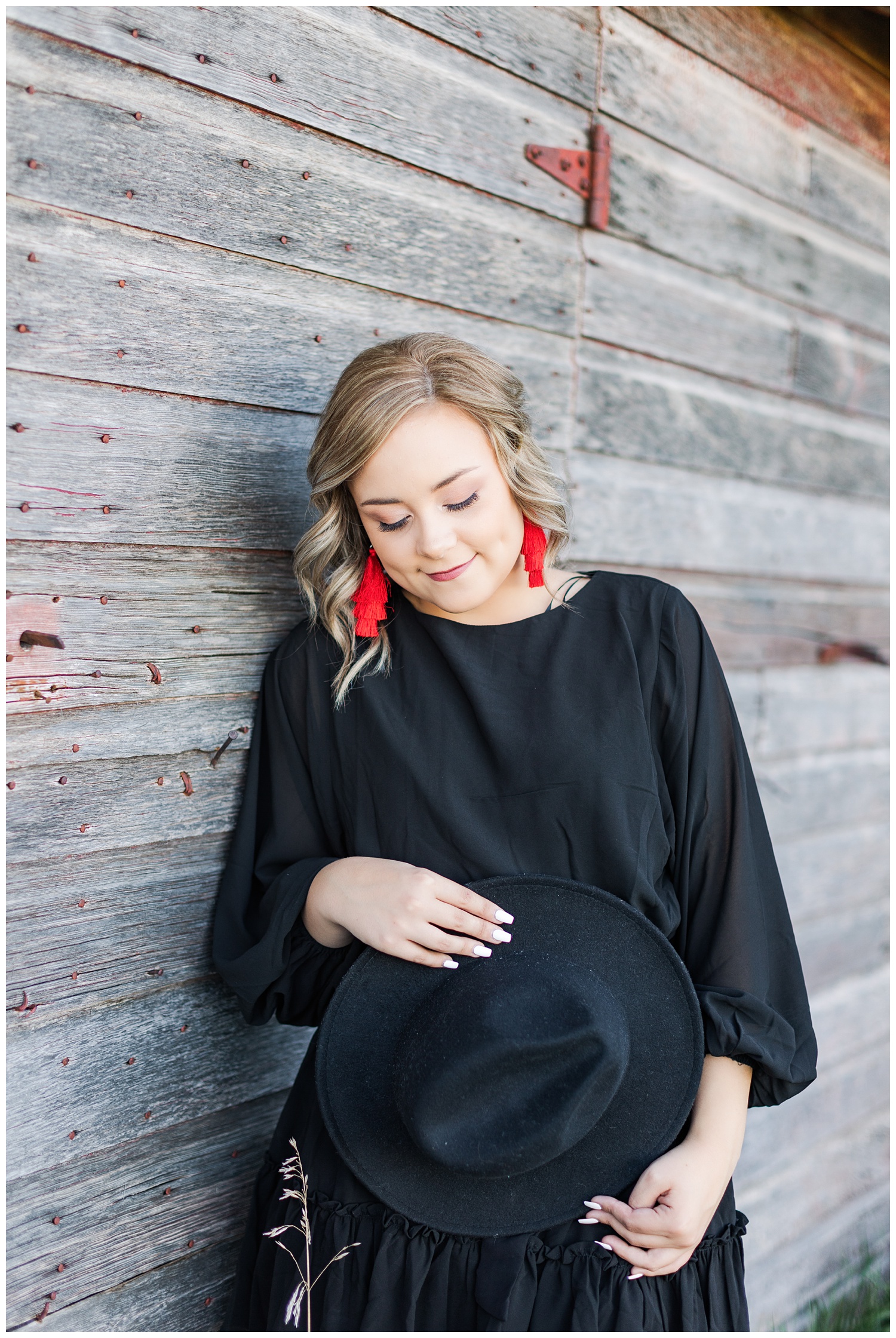 Cloey, wearing a black dress and red, statement tassel earrings, leans up against an old rustic barn while looking down at her hat | CB Studio