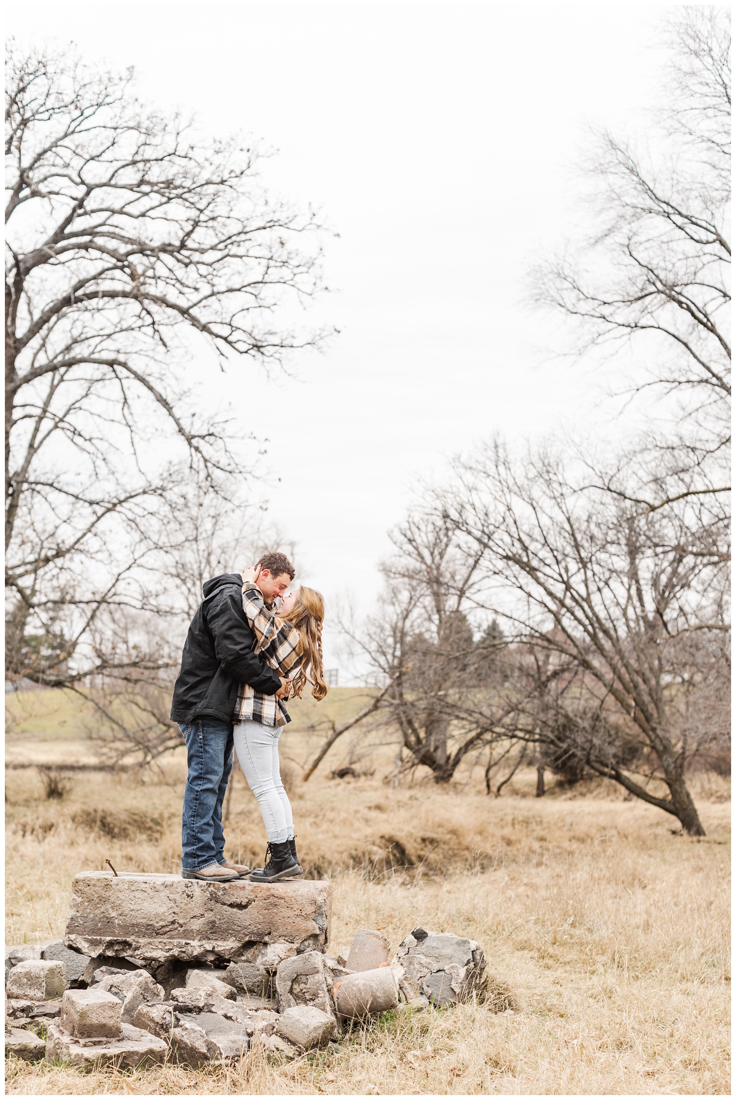 Travis and Abi embrace as they stand on some concrete blocks in a grassy filed in Iowa in the middle of winter | CB Studio