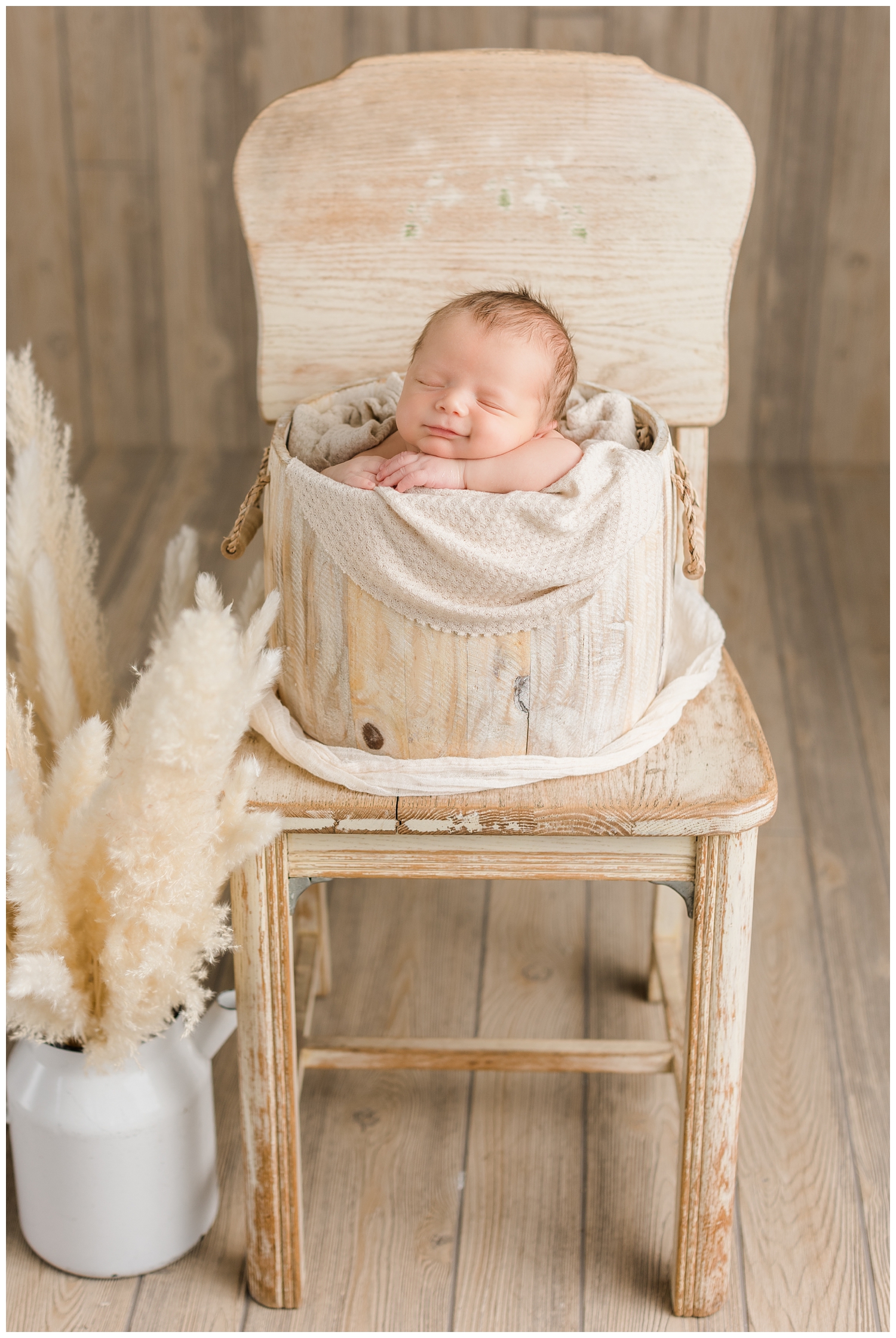 Baby Rhett smiling while nestled in a cream wood bucket on a cream wooden chair with a kettle of pampas grass in a boho theme setup | CB Studio
