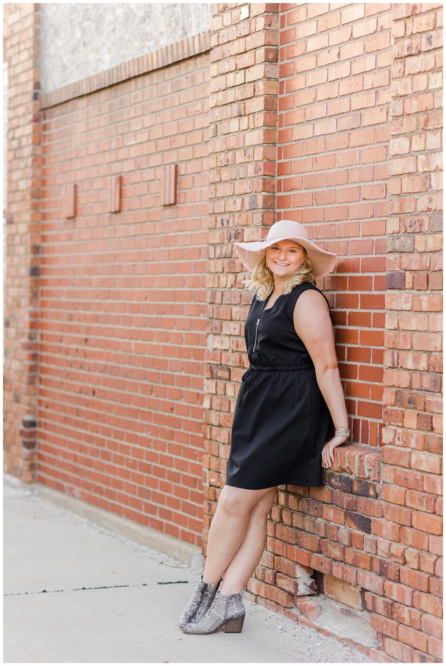 Abby leans against a brink wall in downtown Algona wearing a black dress and a pink sunhat | CB Studio