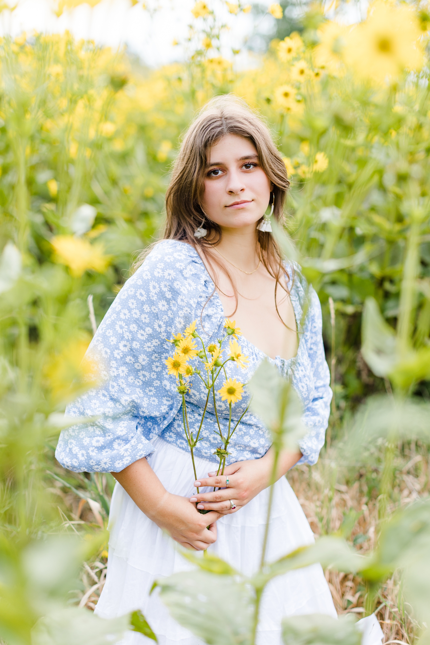 Caitlin stands in a field of yellow wildflowers wearing a baby blue boho pheasant top and a white flowy skirt | CB Studio