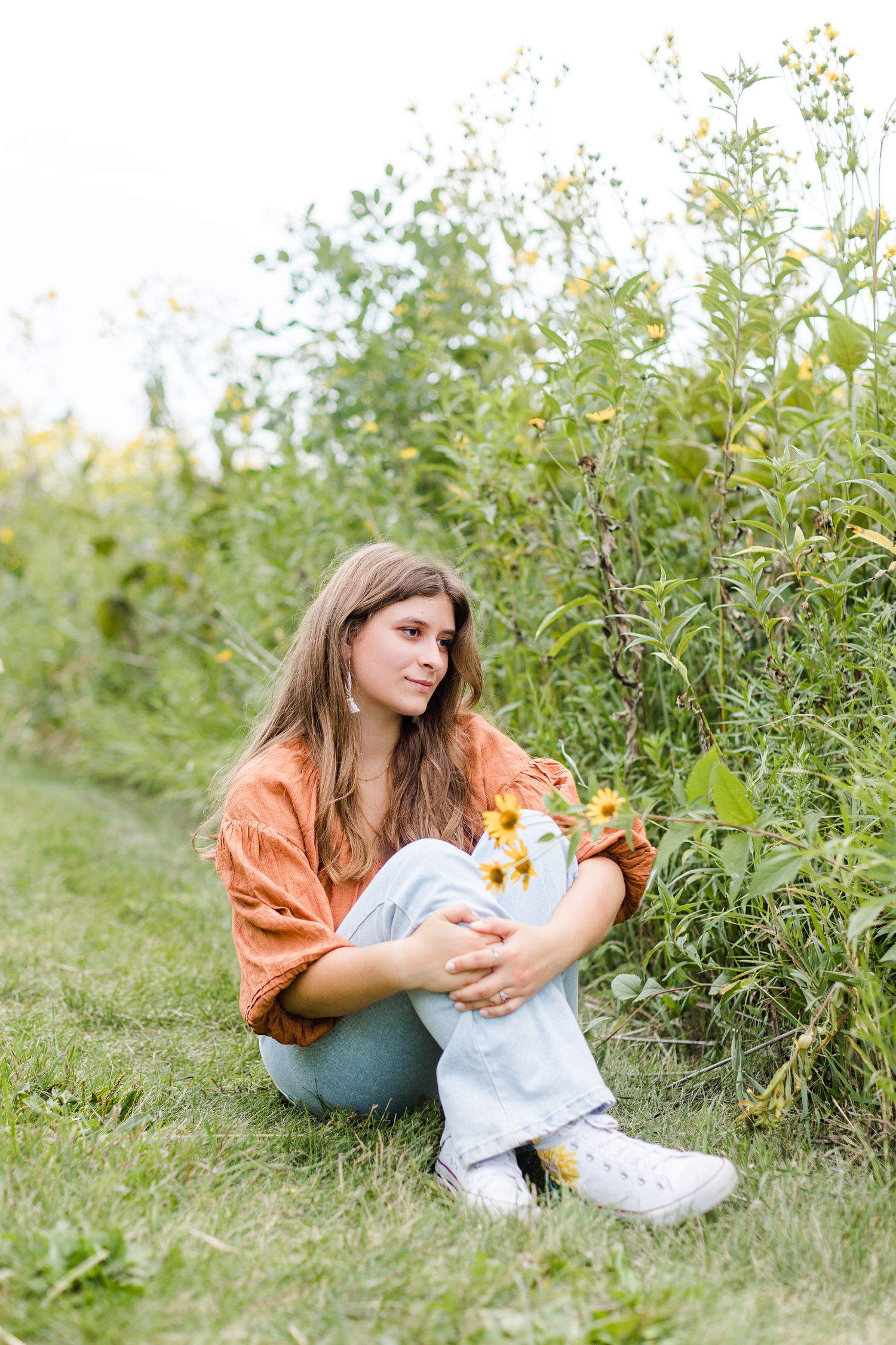 Caitlin sits in a field of yellow wildflowers wearing a boho pheasant top, jeans and daisy sneakers | CB Studio