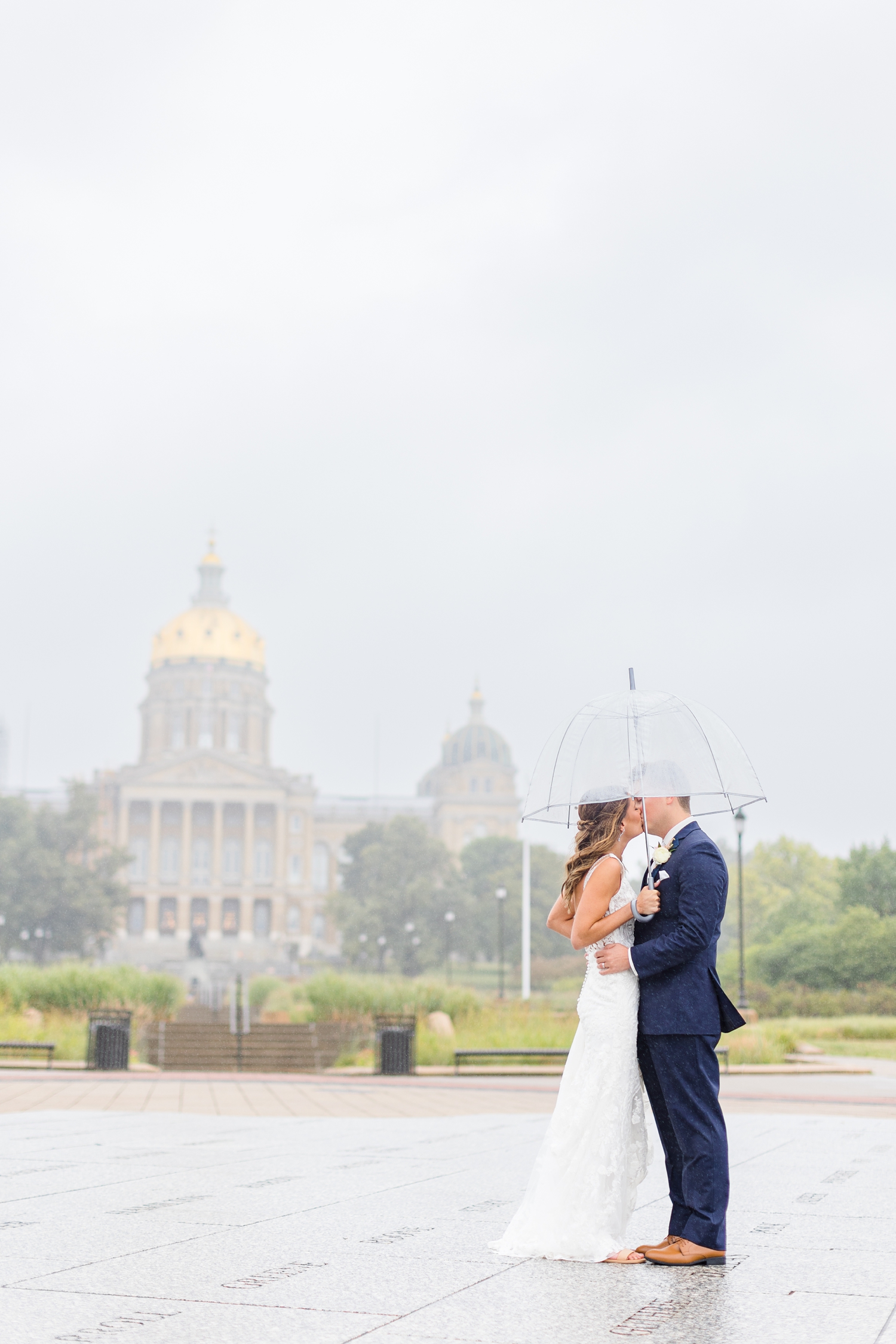 Bride and groom kiss under an umbrella in front of the Iowa capitol while it rains | CB Studio