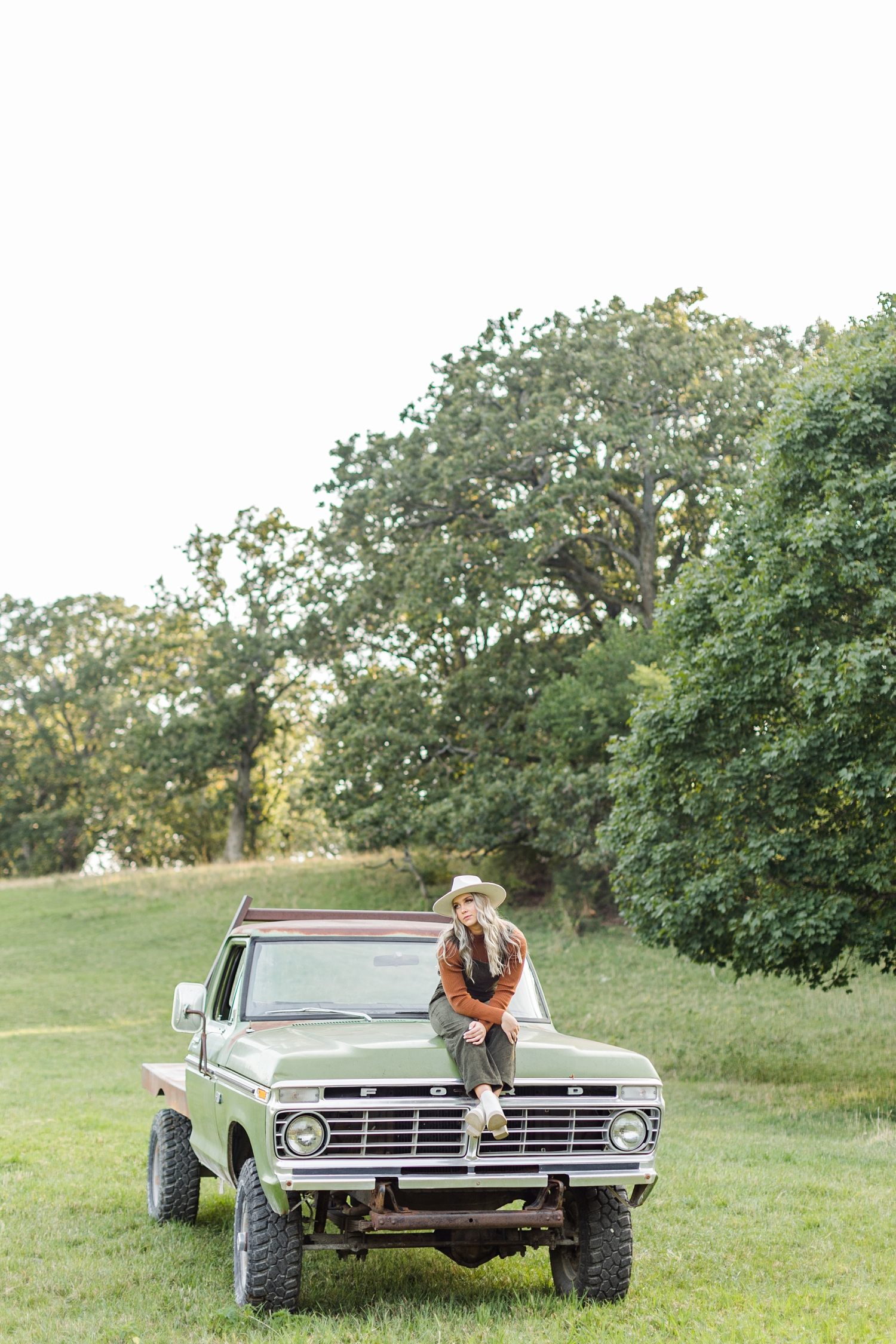 While looking off into the distance, Shelby sits on the hood of a green 72 Ford truck in the middle of a grassy pasture | CB Studio