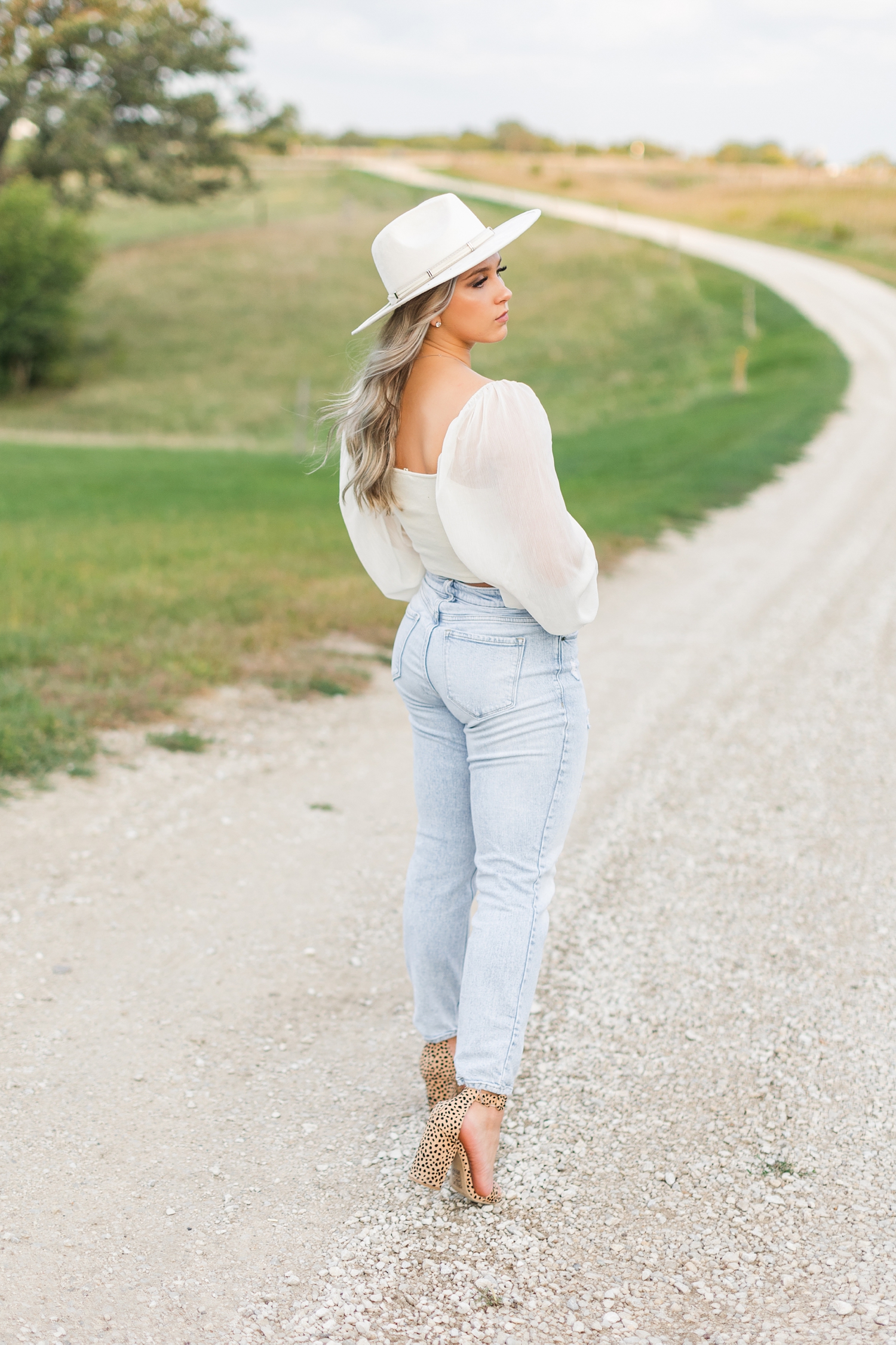 Shelby walks down a gravel road wearing jeans, a cream cropped blouse, wide brim hat and leopard print heels | CB Studio