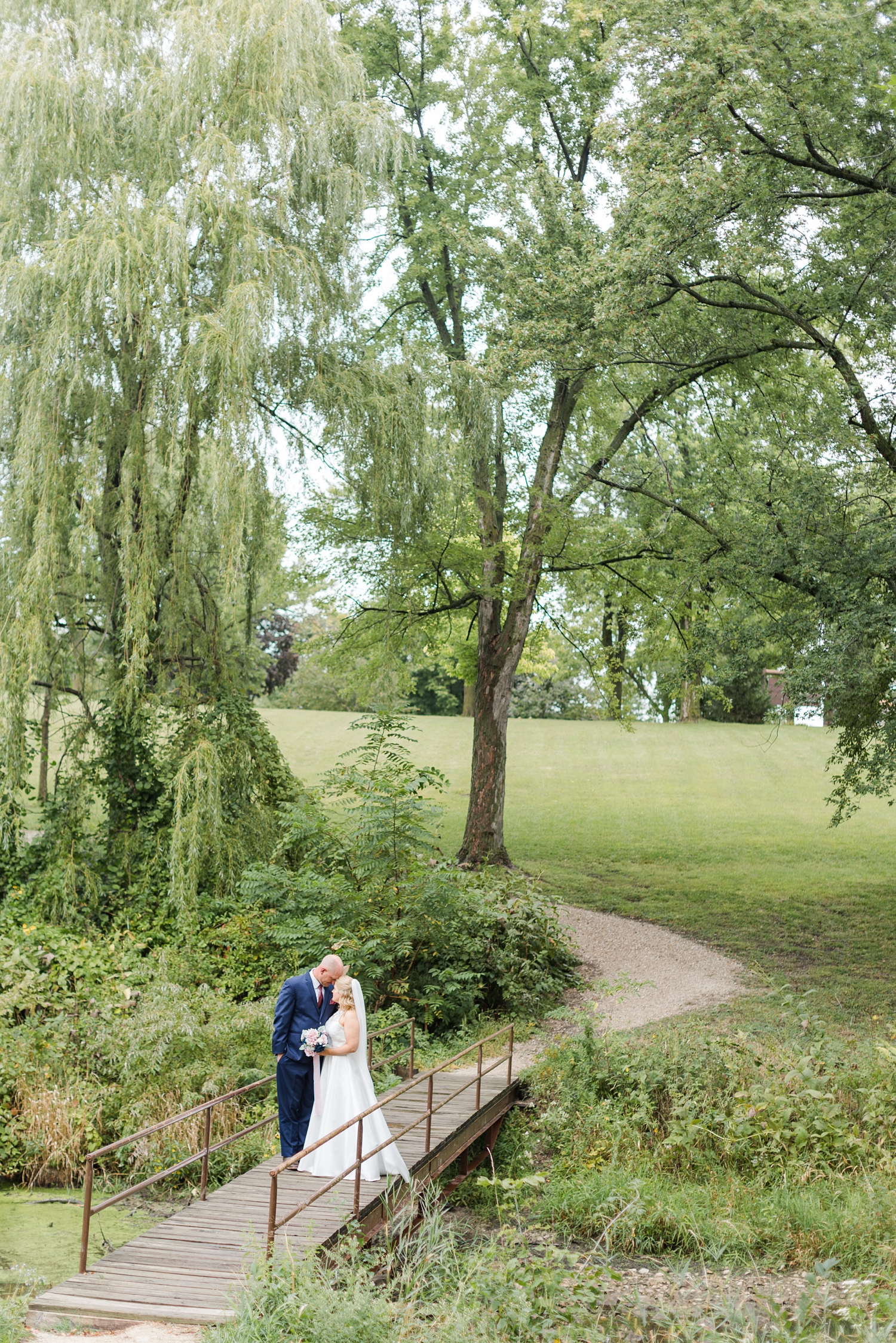 James and Megan embrace on a wooden bridge surrounded by willow trees on their wedding day in Kennedy Park Fort Dodge | CB Studio