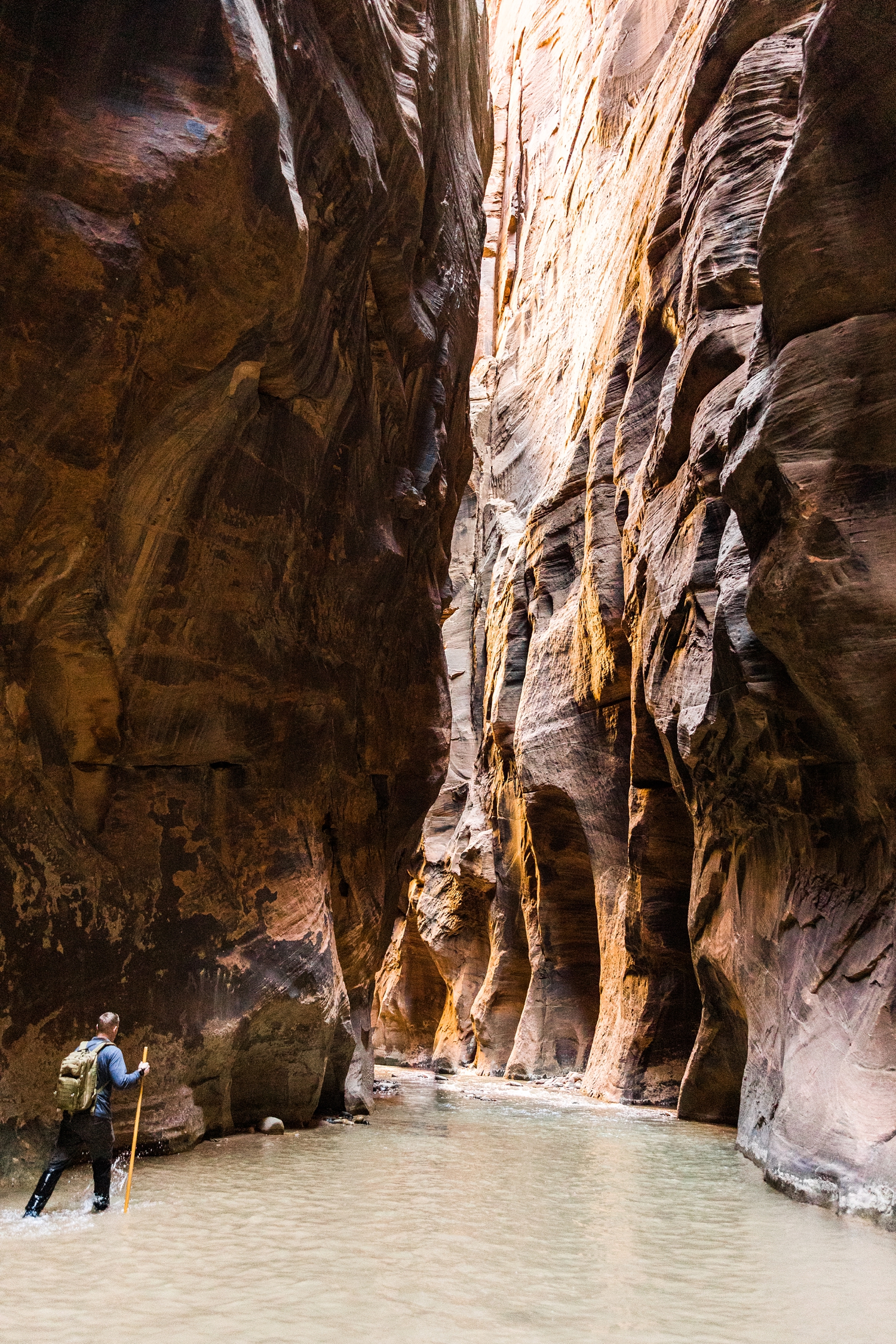 As Joes hikes through the Virgin River, sunrise light shines through the canyon in the Wall Street section of the Narrows hike at Zion National Park | CB Studio