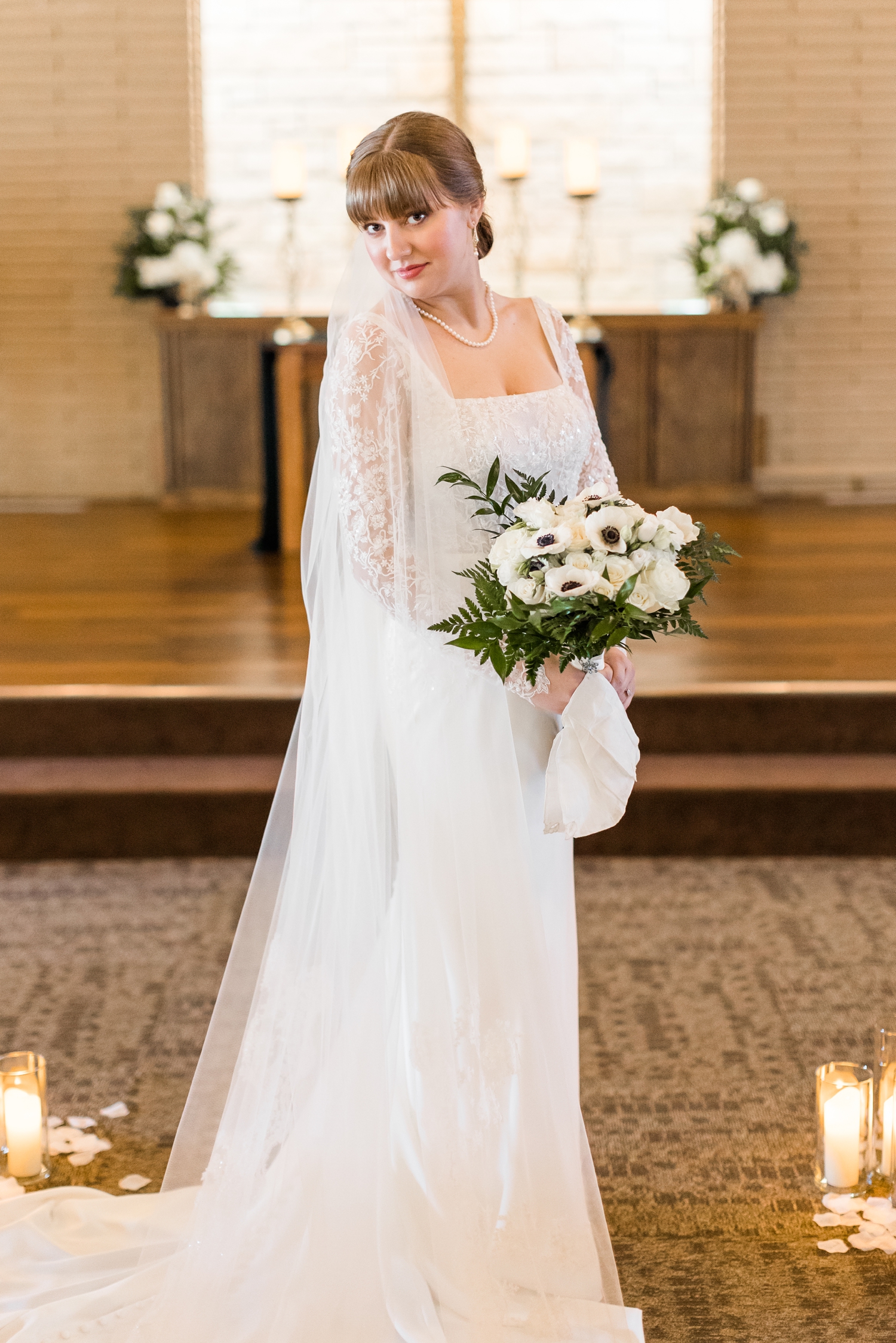 Bride, Madeline, dressed in a custom Stella York long sleeve wedding gown with custom cathedral length veil standing in a wooden church isle surrounded by candlelight holding a white rose and anemone bouquet | CB Studio