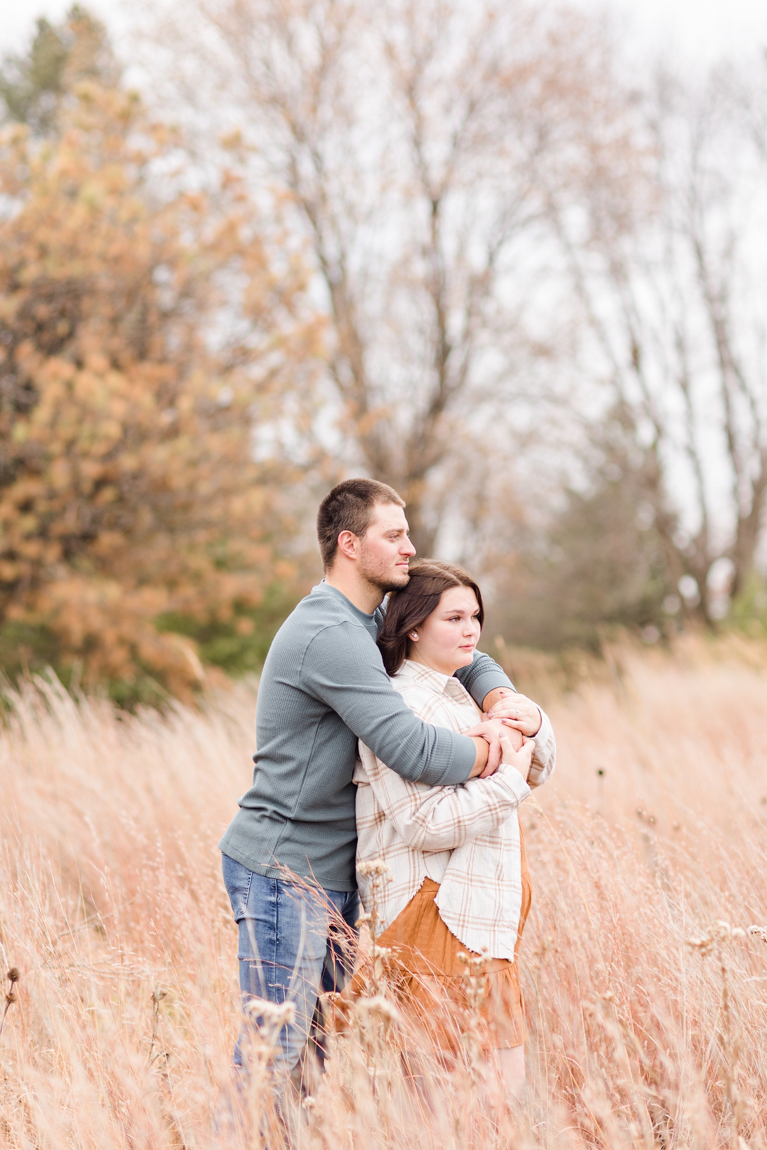 Austin embraces Mary from behind while standing in tall fall grass during their engagement photography session at Kennedy Park in Fort Dodge, IA | CB Studio