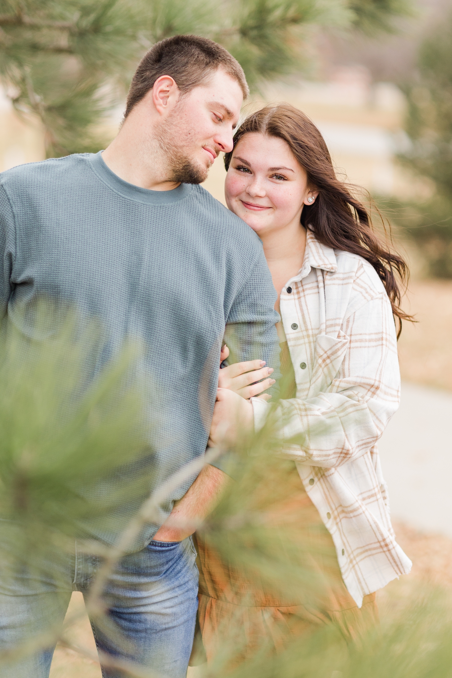Mary hugs Austin's arm from behind soft evergreen trees during their engagement photography session at Kennedy Park in Fort Dodge, IA | CB Studio