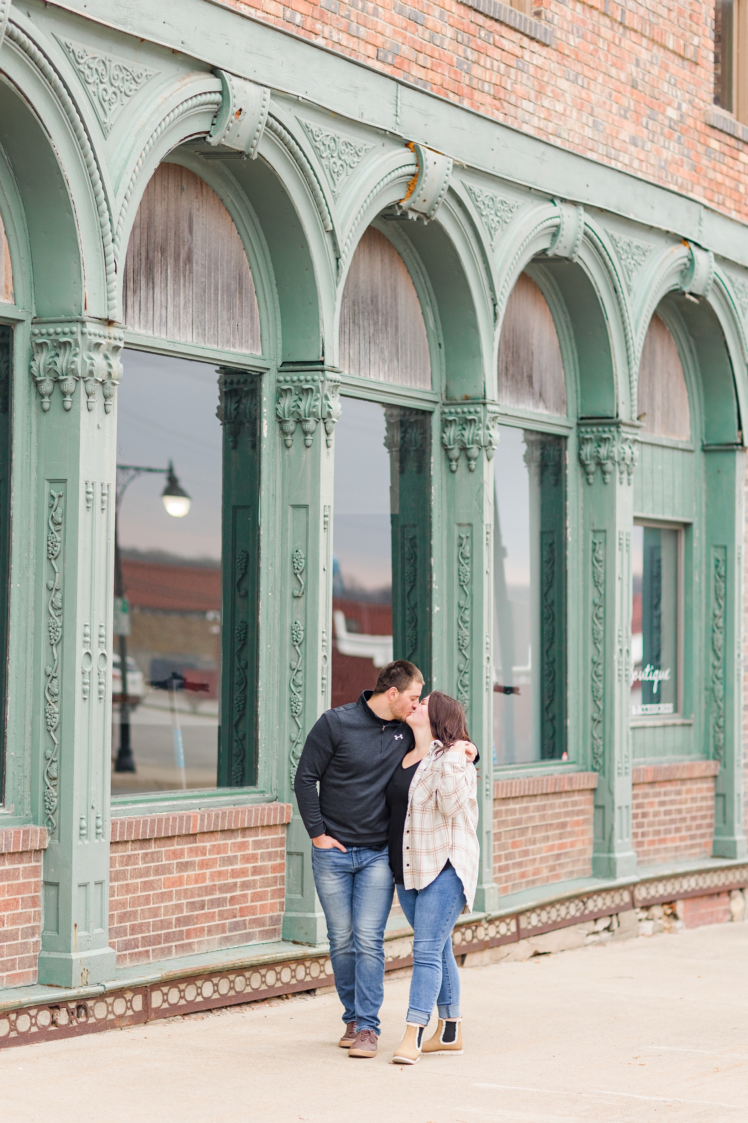 Mary and Austin share a kiss while walking along a historic building with teal architectural arches in downtown Fort Dodge, IA | CB Studio