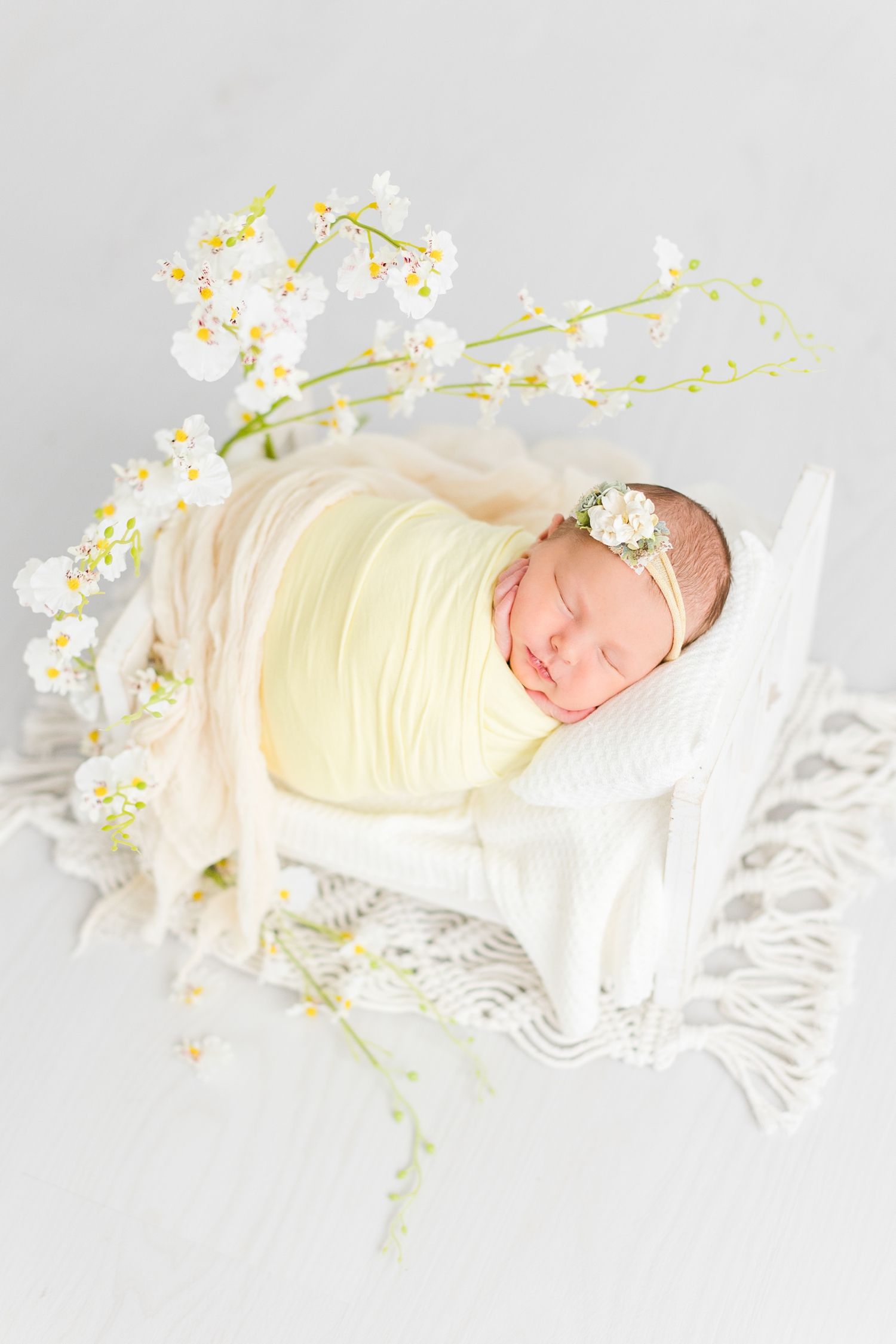 Baby Olivia wrapped in soft yellow laying on a white wooden bed surrounded by white orchid branches | CB Studio