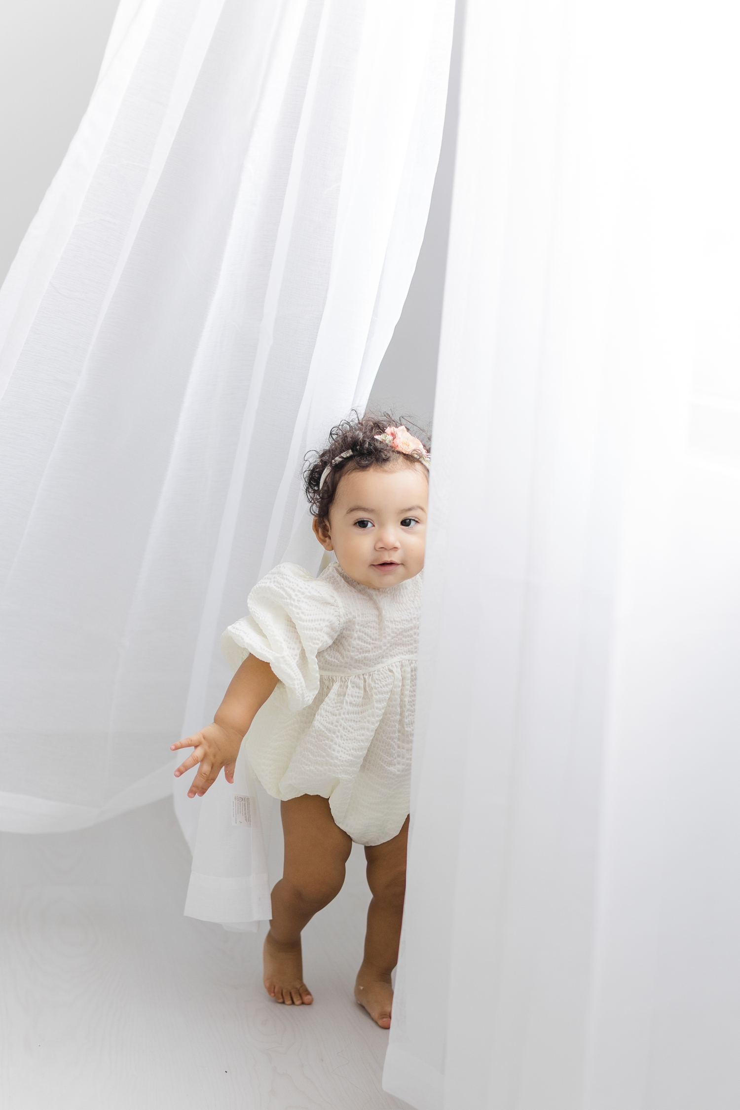 Baby Zoey dressed in a white bubble romper playing joyfully in white flowy curtains | CB Studio