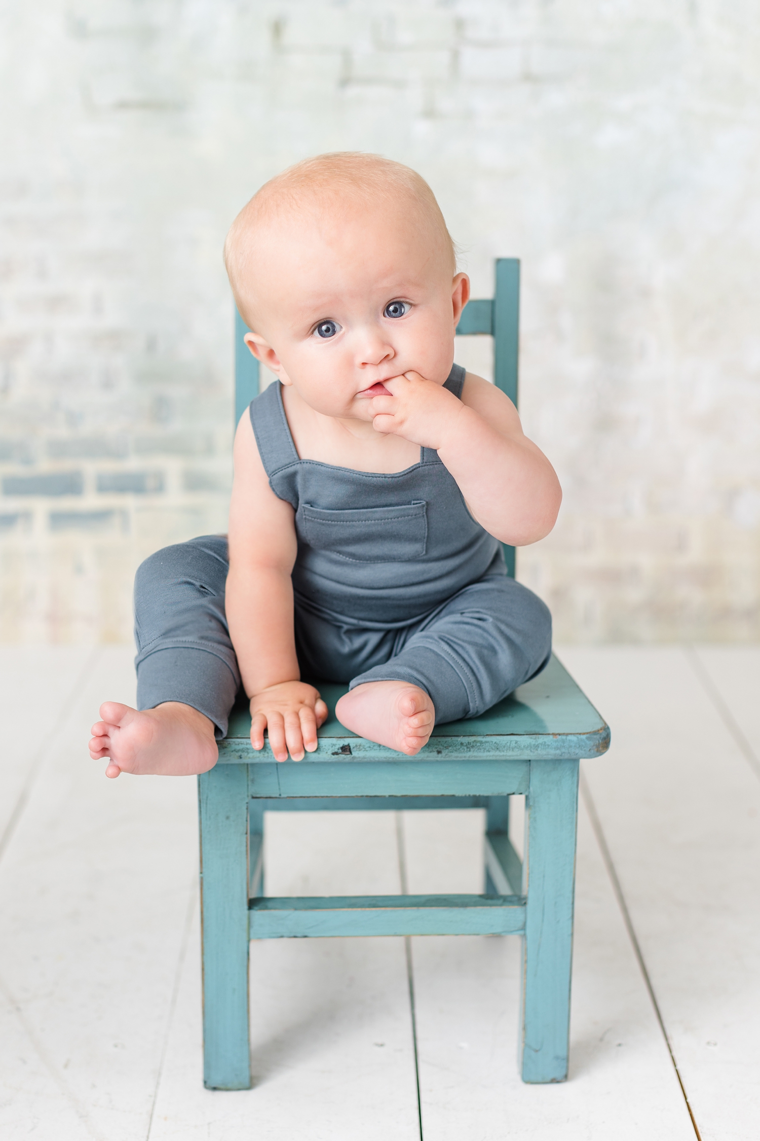 Baby Hudsyn sits in a blue chair wearing blue overalls, sucking on his fingers | CB Studio