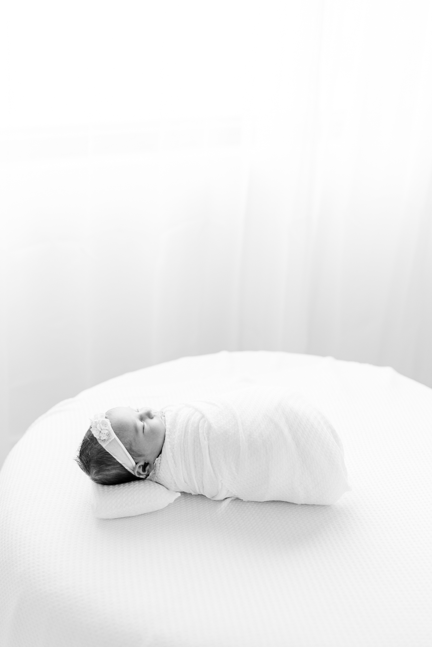 Baby Ivy wrapped in white laying on a white ottoman with white curtains and window in the background | CB Studio