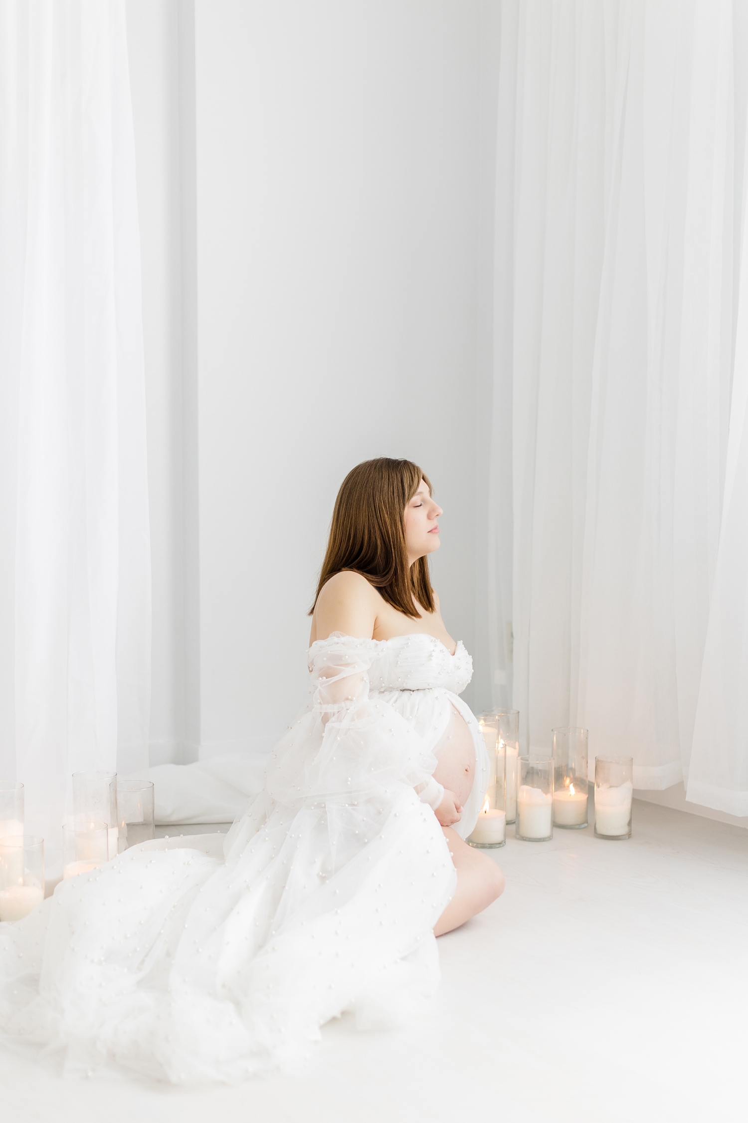 Madeline sits while gently holding her baby belly, wearing a white tulle maternity gown covered in pearls and surrounded by white draping clothes and candlelight | CB Studio Photography