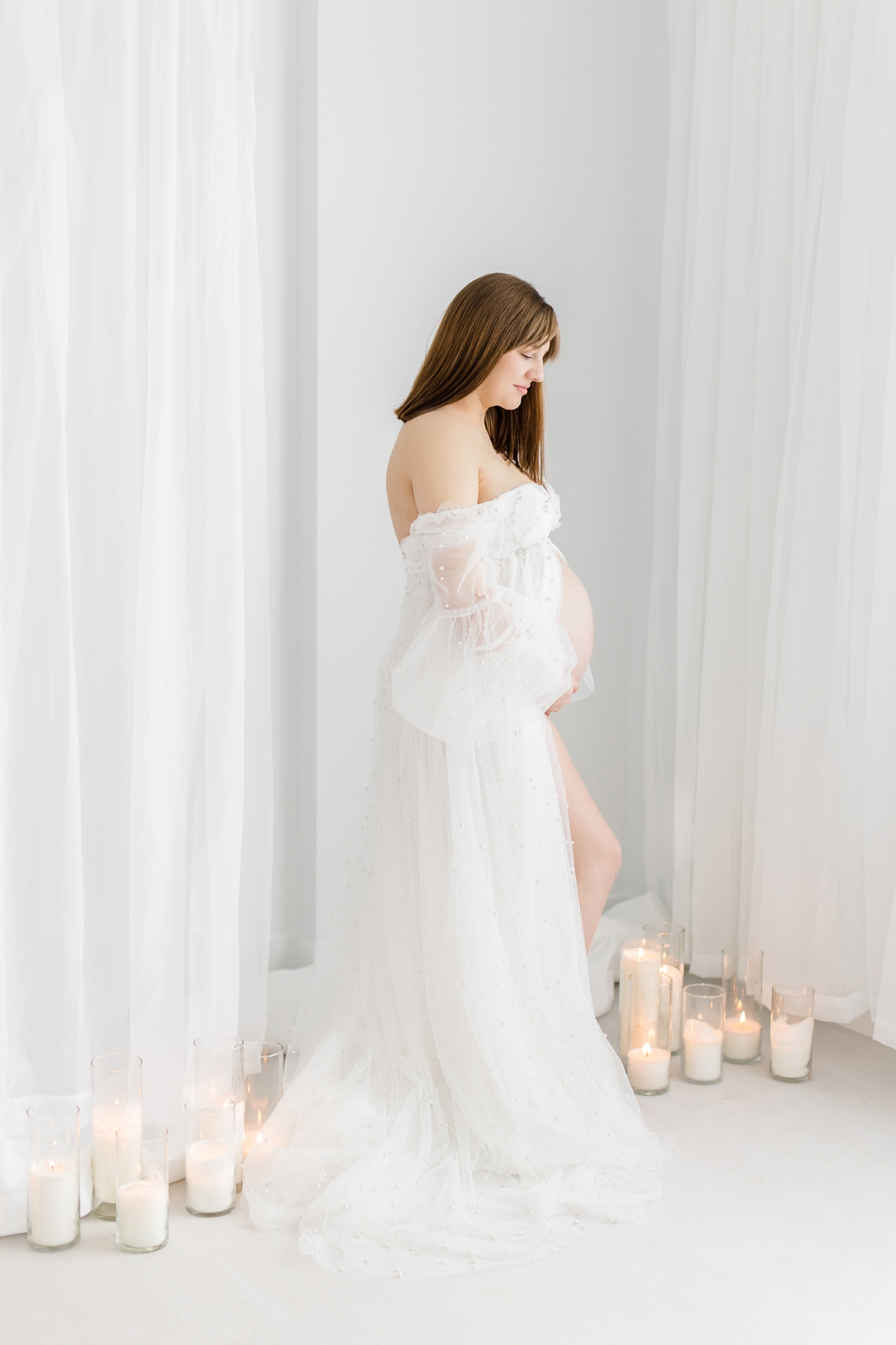 Madeline gently holds her baby belly while wearing a white tulle maternity gown covered in pearls and surrounded by white draping sheers and candle light across the floor | CB Studio Photography