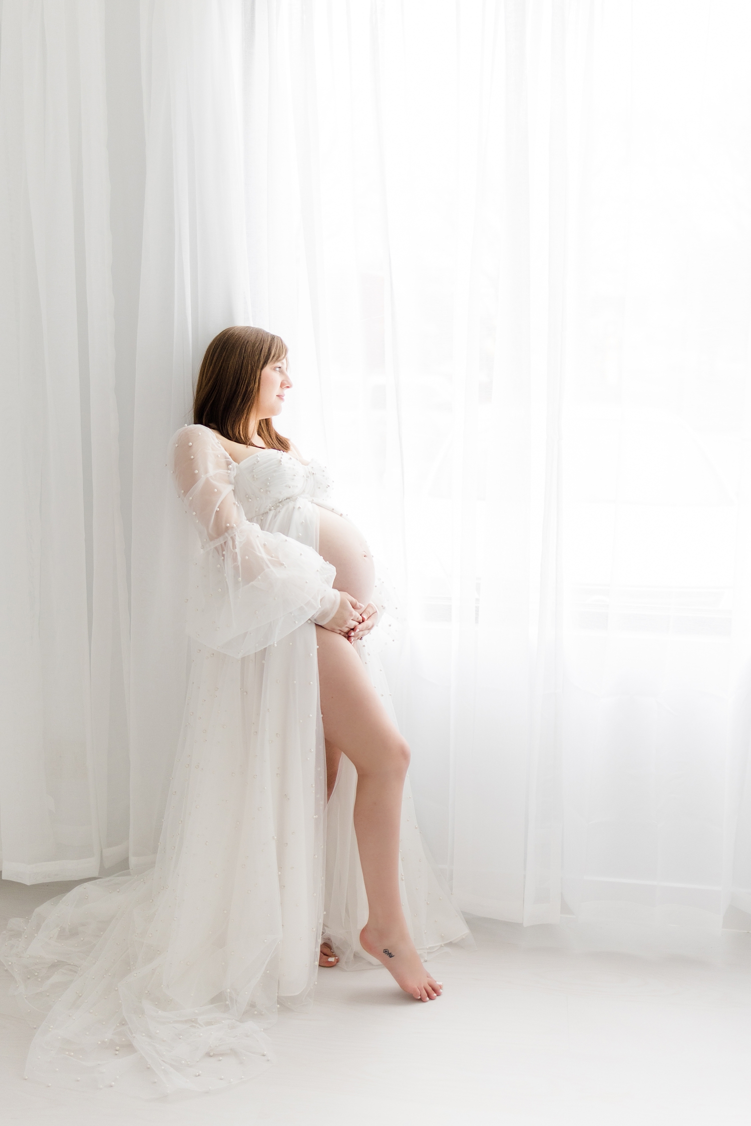Madeline gently holds her baby belly while staring out the window wearing a white tulle maternity gown covered in pearls | CB Studio Photography