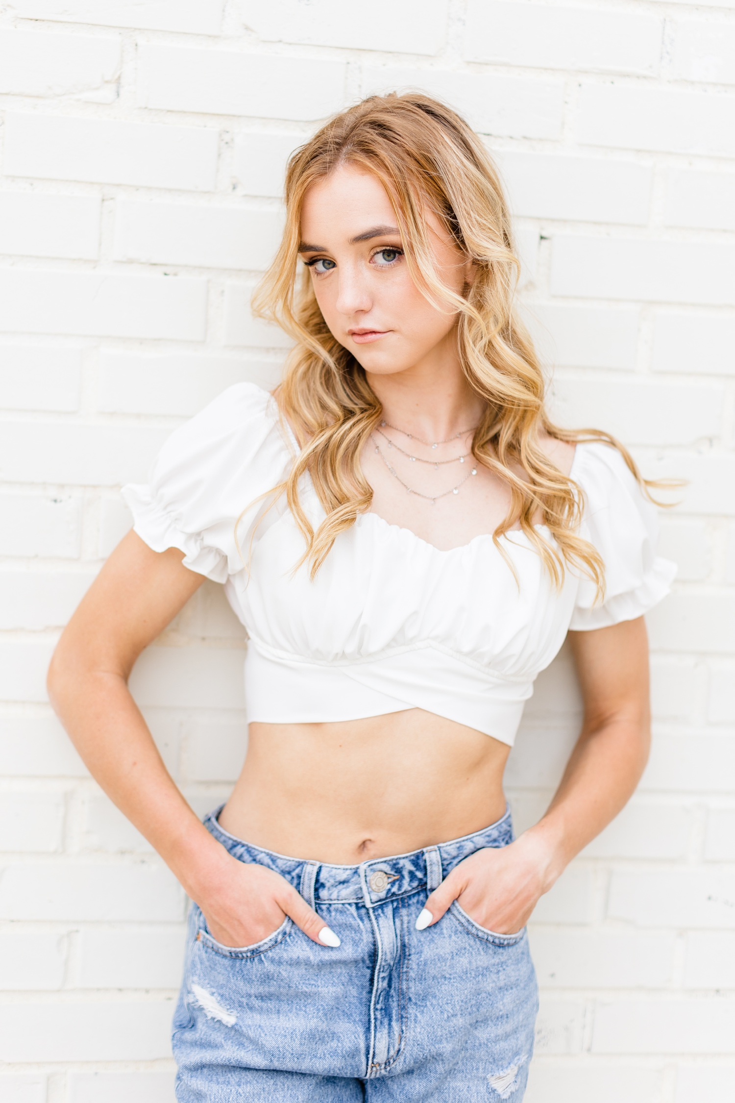 Reagan, Bishop Garrigan Class of 2024, leans against a white-wash brick building, wearing jeans with a white crop top with ruffled sleeves. | CB Studio