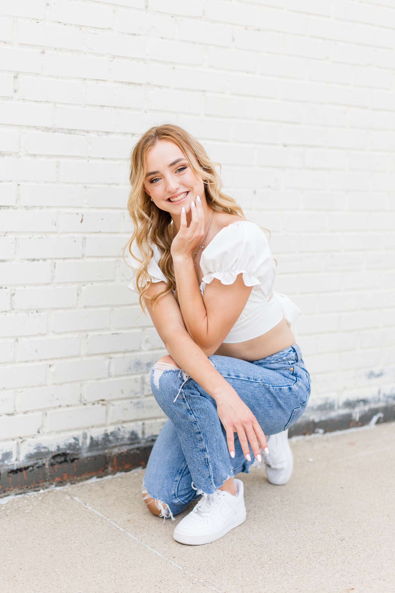 Reagan, Bishop Garrigan Class of 2024, squats down by a white-wash brick building, wearing jeans with a white crop top with ruffled sleeves. | CB Studio