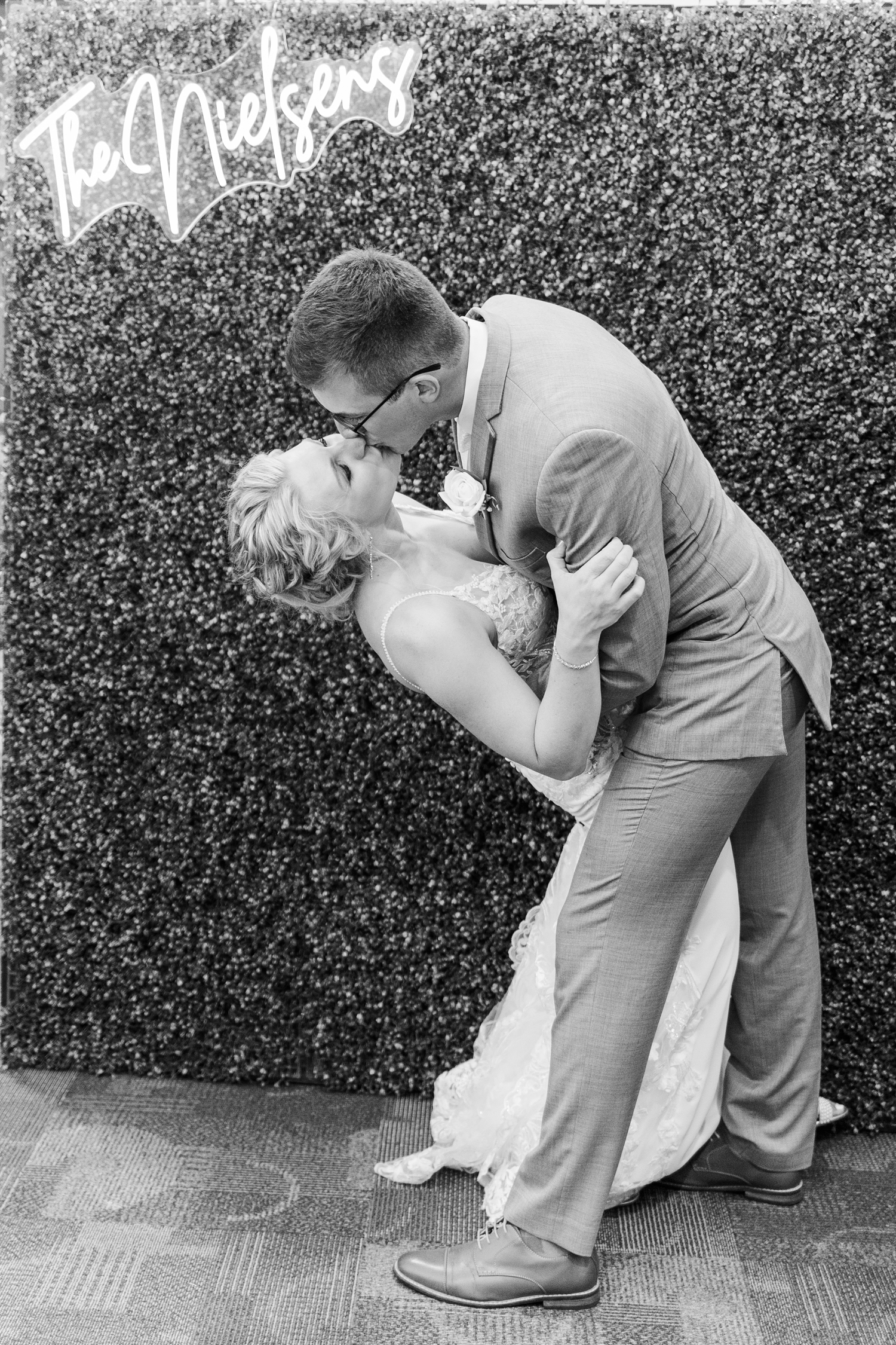 Quinton dips is new bride, Alli, for a kiss in front of a boxwood background complete with a neon sign that reads "The Nielsens" | CB Studio