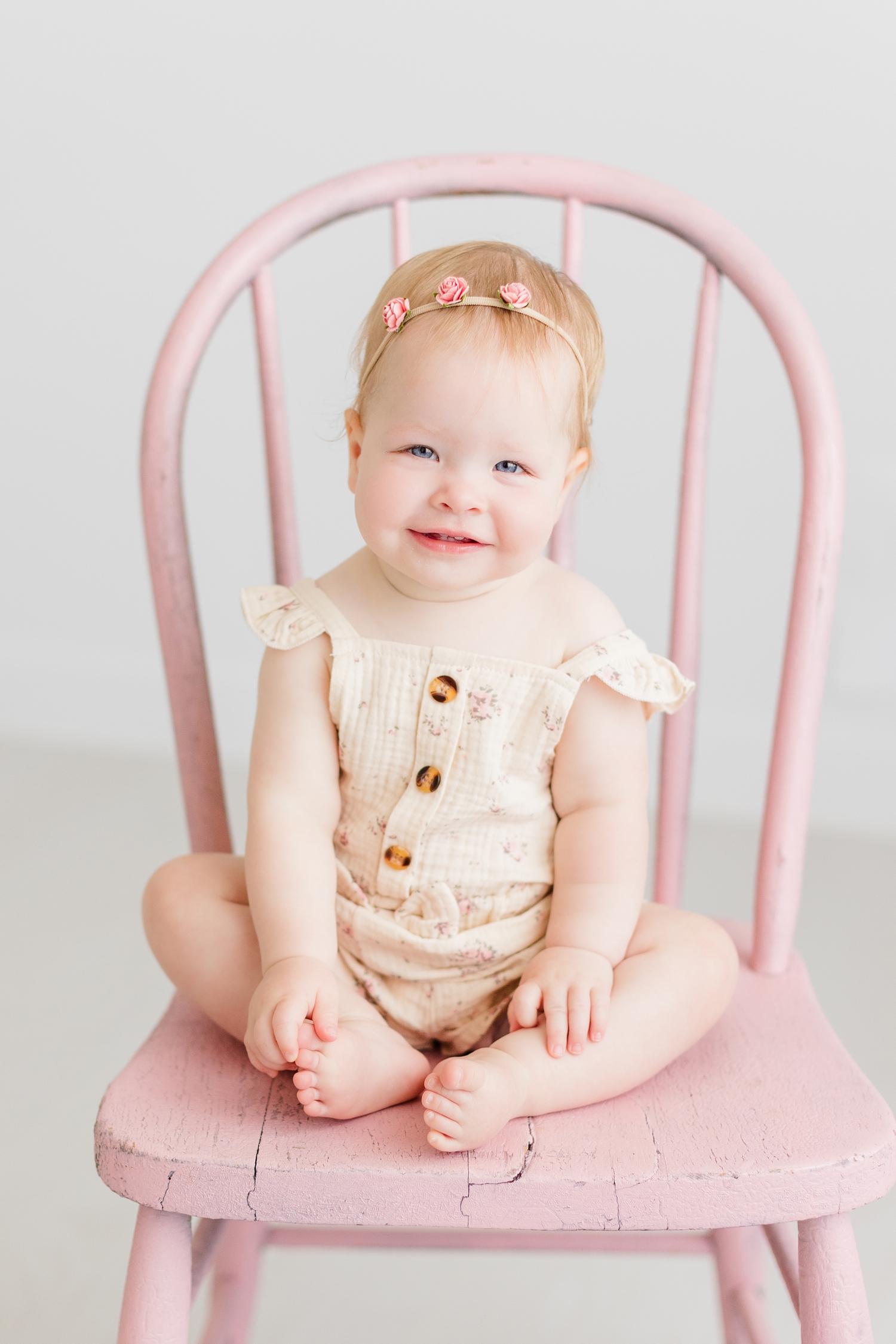 Baby Shae, wearing a cream gauze fabric romper covered in vintage roses sits in a pink wooden chair in an all white room | CB Studio