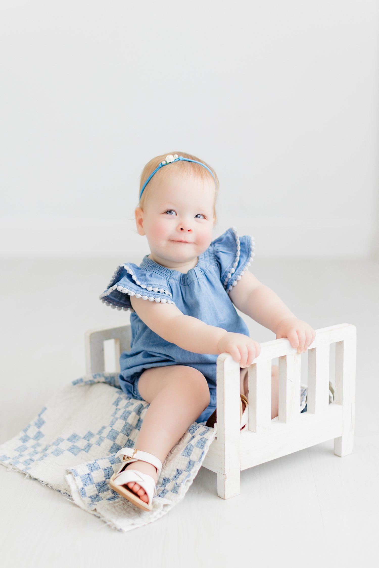 Baby Shae, wearing a denim blue colored romper sits in a white, wooden doll bed draped with a white and blue quilt | CB Studio