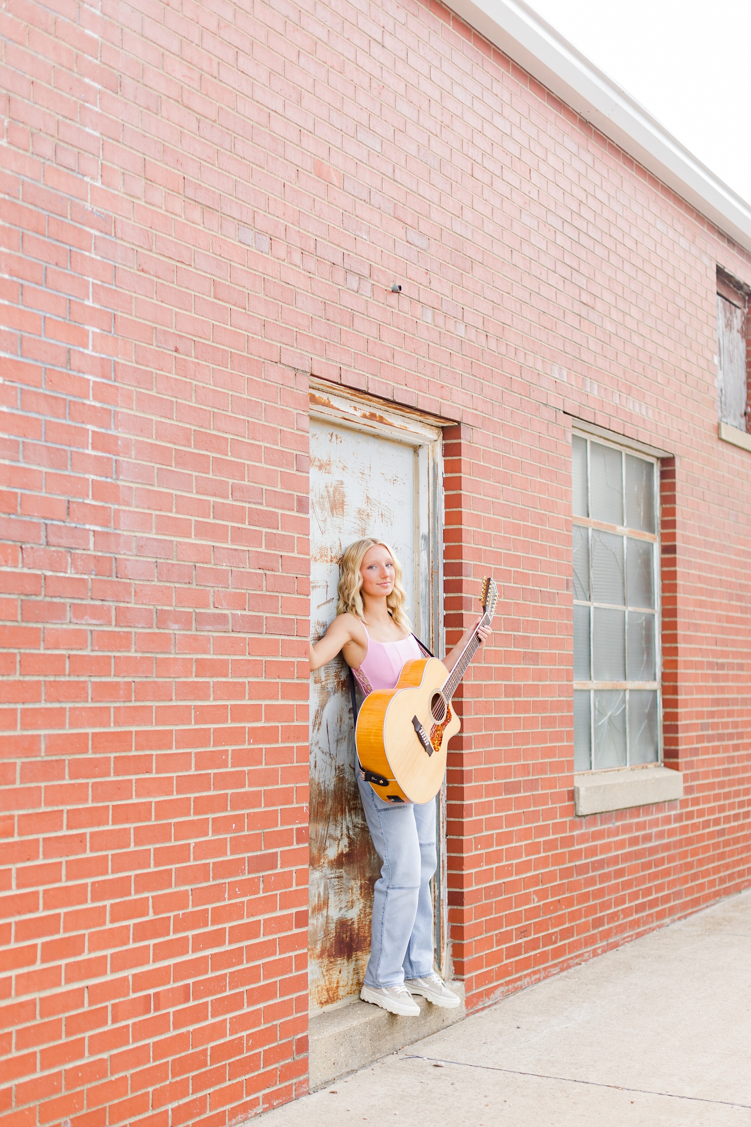 Avery stands in the doorway of an old brick warehouse holding her guitar in downtown Algona, IA | CB Studio