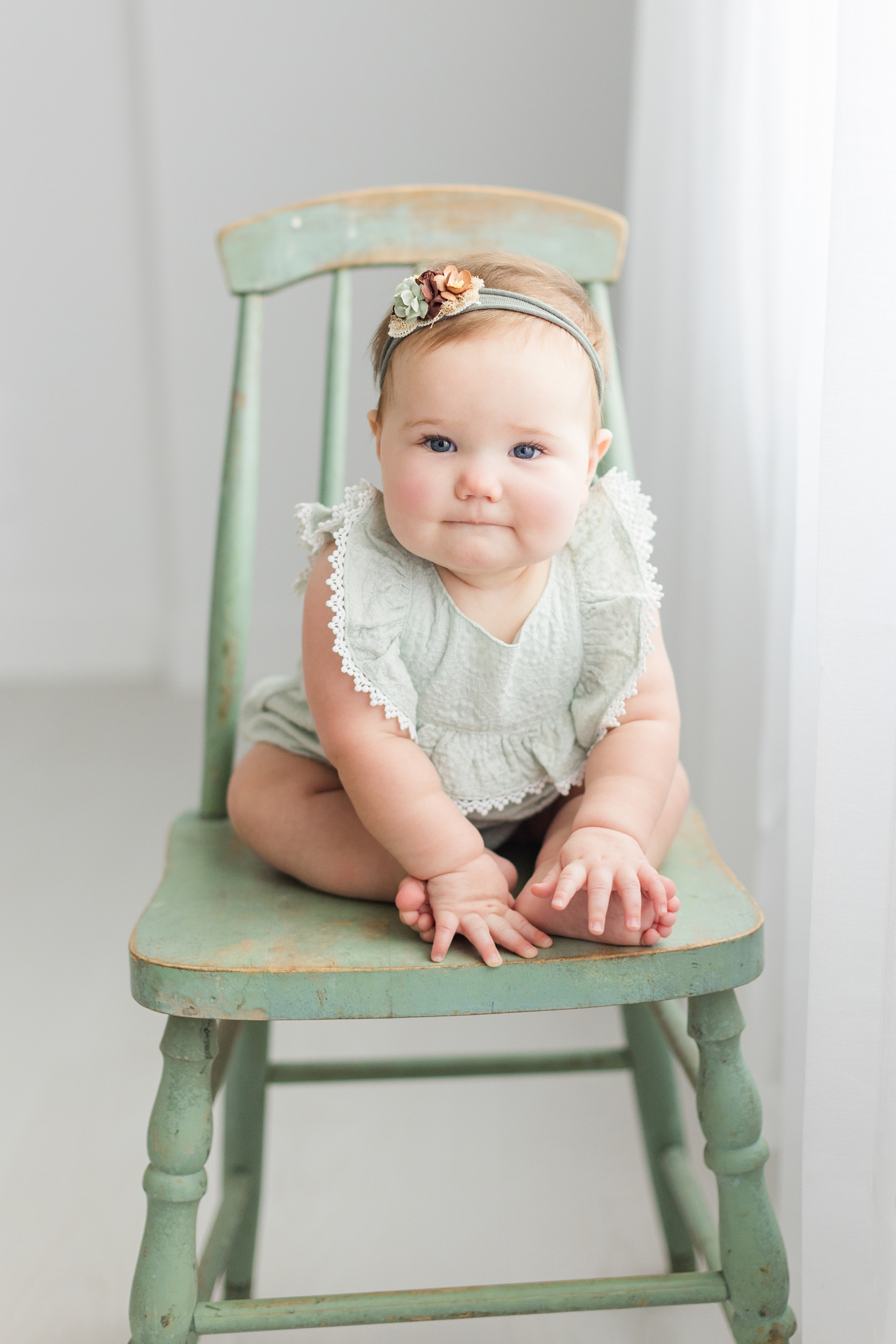Ivy sits on a vintage sage green chair next to long, white, flowy curtains, wearing a sage green romper and sage green headband with fall colored flowers | CB Studio